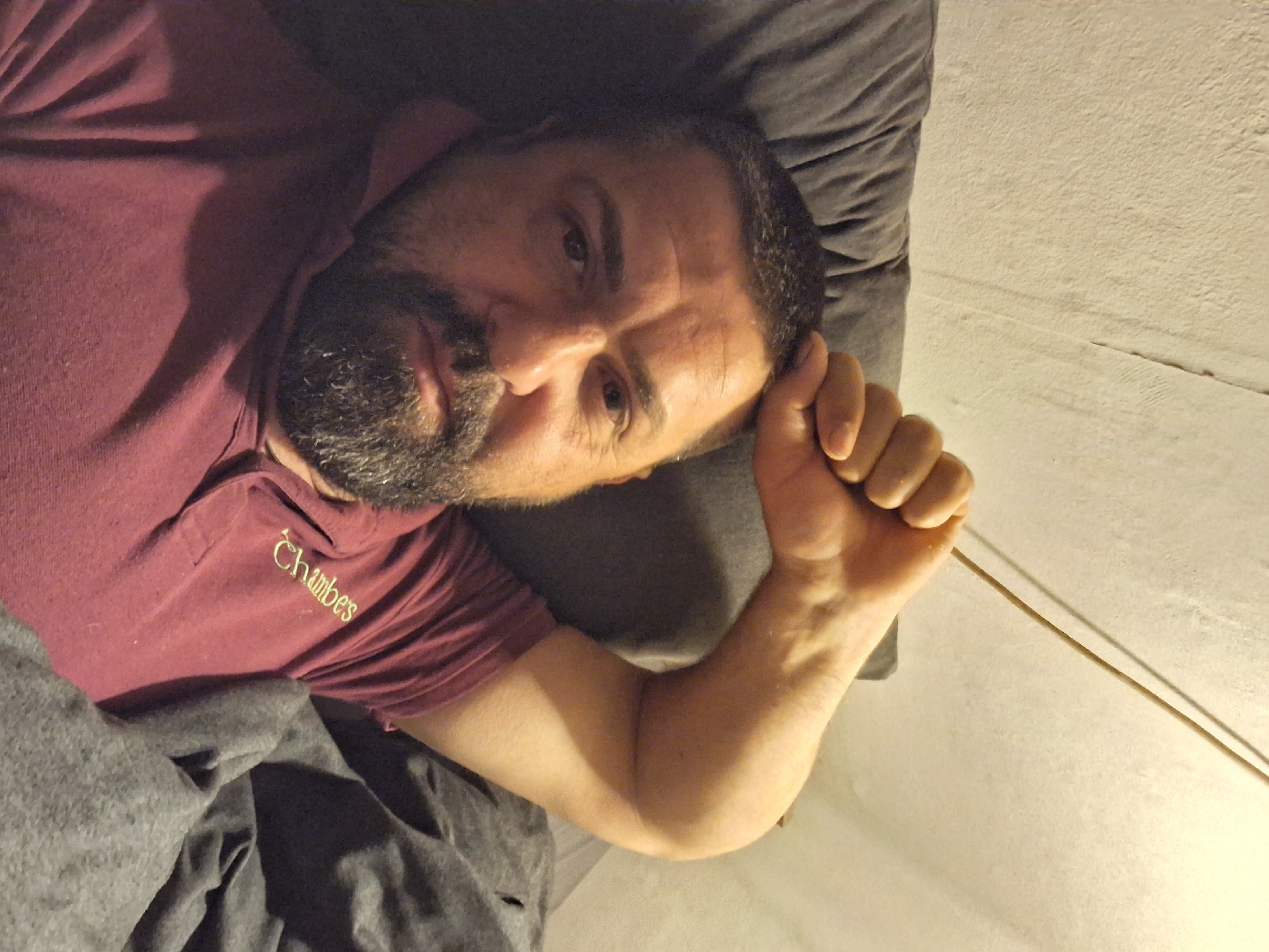 A man with a beard lies on a pillow in bed. He is wearing a burgundy polo shirt with the name "Chambers" embroidered on it. He is covered by a gray blanket. His right arm is raised and his hand is clenched into a fist. The location is not possible to determine based on the image.