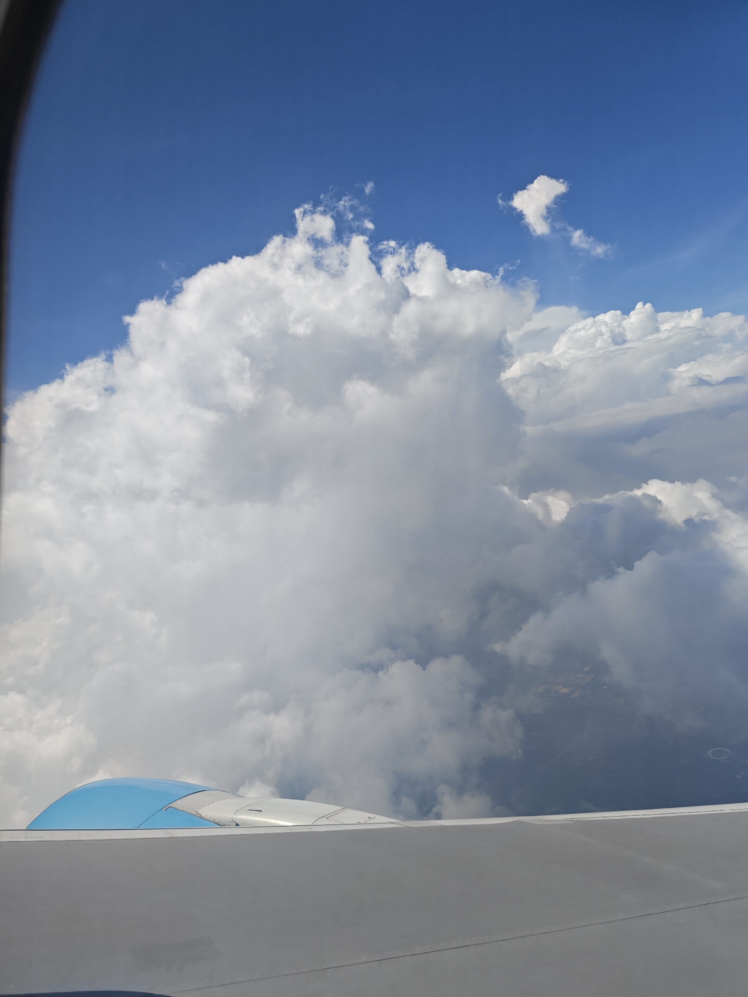 A view from an airplane window shows a vast expanse of white clouds against a bright blue sky. The wing of the airplane is visible in the foreground, with a portion of the blue engine casing also in view. Below the clouds, a glimpse of the ground can be seen, appearing as a dark, textured landscape, possibly indicating the area of Bissar Akbarpur, India. The image captures a moment of flight, with the vastness of the sky and the earth stretching out below.