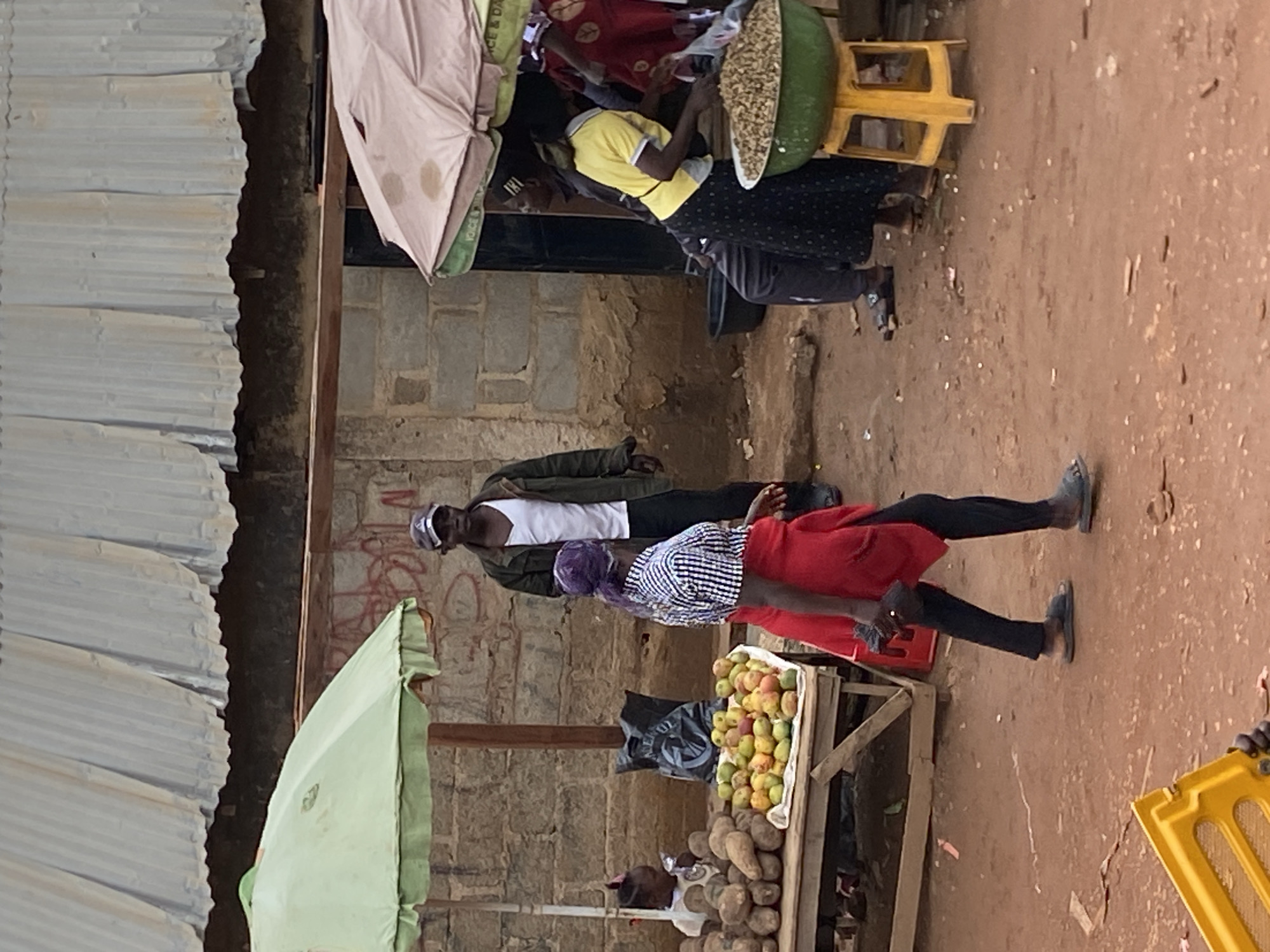 The image depicts an outdoor scene in Bukuru, Nigeria. People are present, engaging in commerce around informal market stalls. One stall displays fruits and root vegetables. Individuals are either standing or seated near these stalls. Two umbrellas offer shade to vendors and customers. The environment is characterized by a dusty ground, and buildings of brick and corrugated metal.
