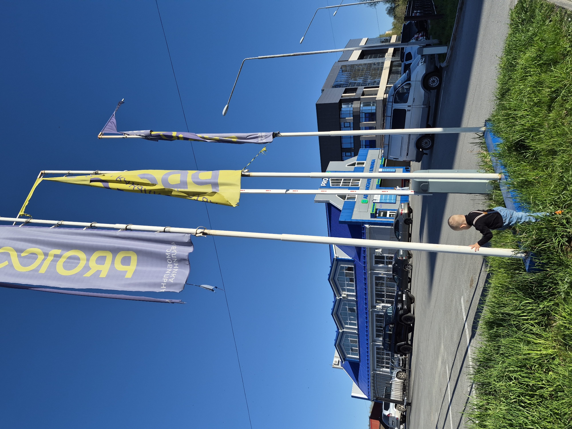 A low-angle shot shows a clear blue sky above an urban scene in Tobolsk, Russia. A young boy leans on a white pole next to a grassy area, wearing jeans and a black top. Behind the boy is a paved parking lot lined with vehicles, including a silver van. In the background, there is a modern building with a blue and grey facade, as well as street lights. Three vertical poles extend upwards into the sky, each with a banner attached. The banners are a mix of yellow and blue colors, displaying Russian text.