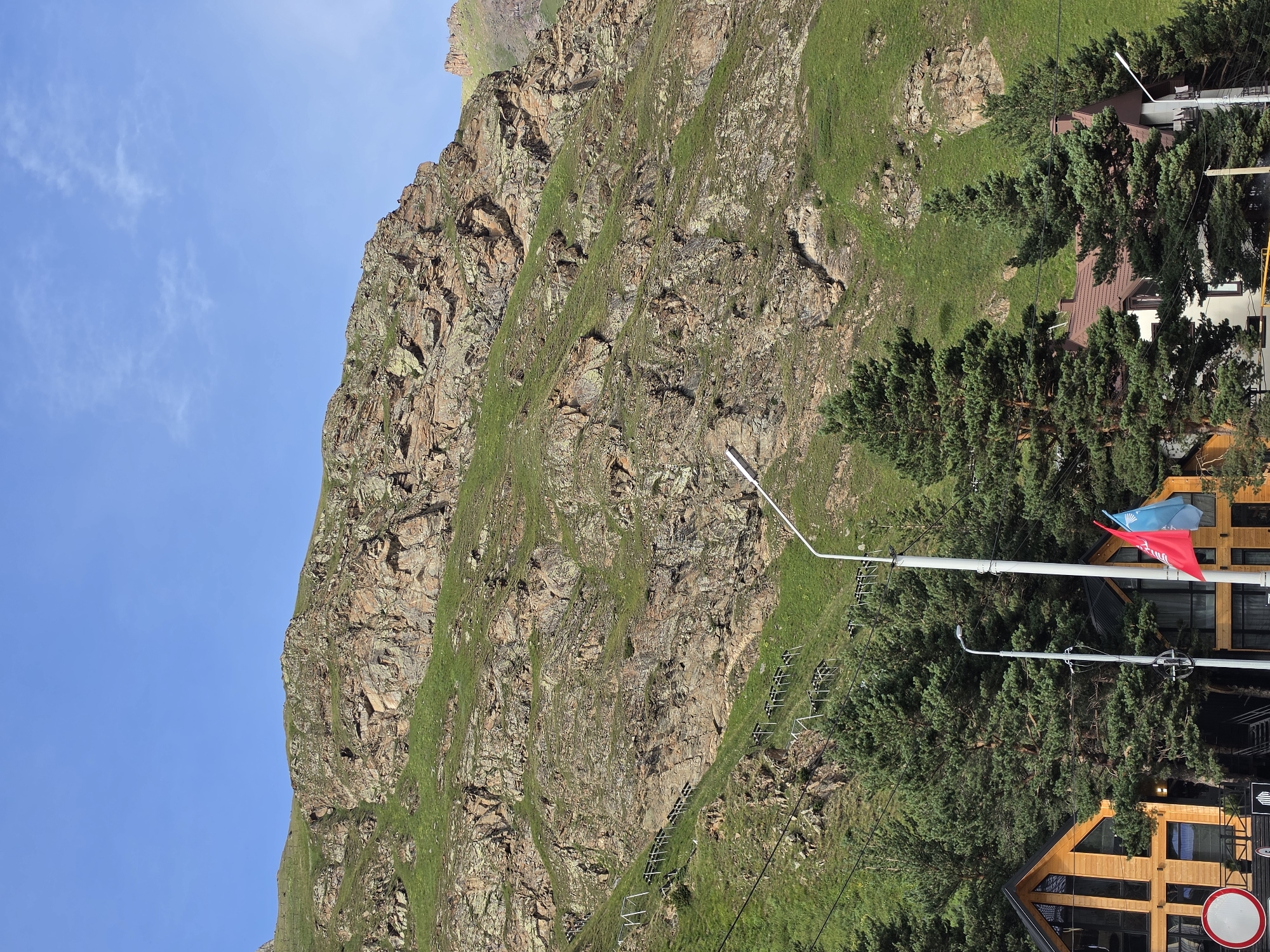 The image depicts a mountainous landscape in Terskol, Russia, under a clear blue sky. A steep, rocky mountain face covered in patches of green grass dominates the left side of the frame. In the lower right quadrant, several modern wooden buildings are nestled amongst tall evergreen trees. Two flags, one red and one blue, are visible flying from a pole attached to one of the buildings. A street lamp is also present on a pole. The scene appears to be set during the daytime, with bright sunlight casting shadows. There are no people visible in the image. The overall impression is of a serene, natural environment with human habitation integrated into the mountainous terrain.