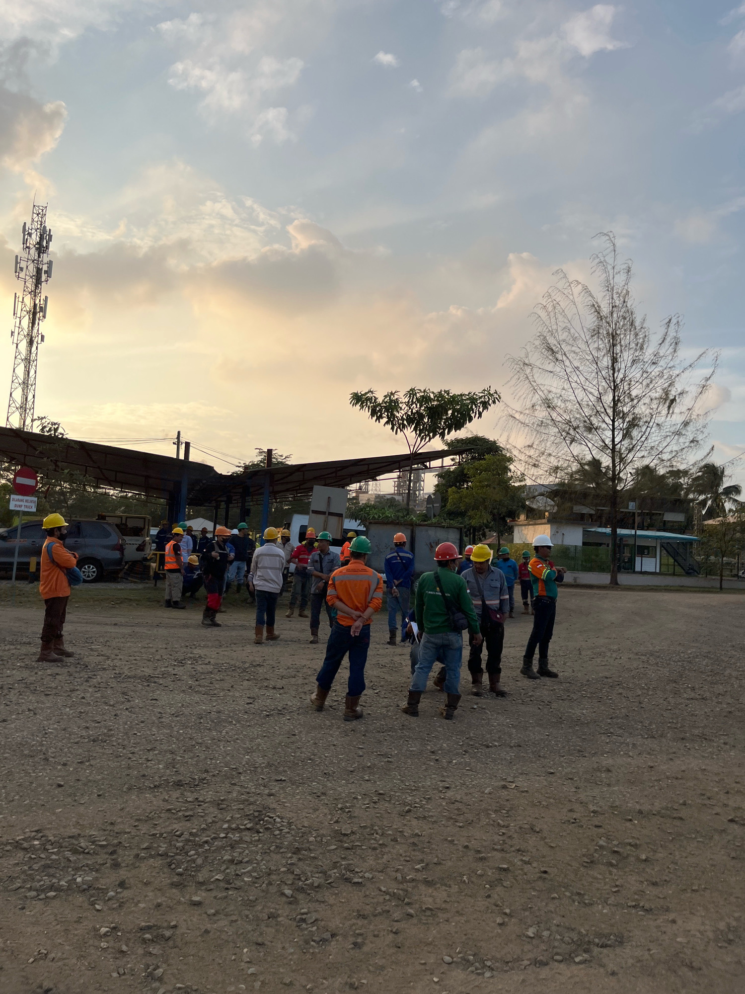 A group of approximately twenty individuals, all wearing hard hats and safety vests, are gathered in an outdoor, gravel-covered area. The scene appears to be a construction site or industrial facility. The sky is filled with clouds, tinged with the warm colors of either sunrise or sunset, indicating it is early morning or late evening.
In the background, a tall telecommunications tower stands to the left of the frame. Several vehicles, including a car and what appears to be a small bus or van, are parked under a covered structure. Further in the background, there are buildings and trees, suggesting an urban or semi-urban environment. The ground is uneven and covered with small stones and dirt.
The individuals are not engaged in any specific activity, but some appear to be listening or observing. The overall atmosphere is one of a work-related gathering. The location is identified as Unknown, Indonesia, based on the context provided.