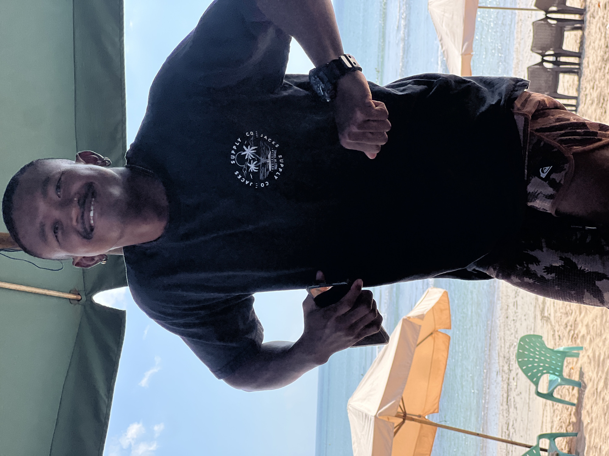 A man with a mustache and earrings stands on a beach in Kuta, Indonesia, smiling at the camera. He is wearing a black t-shirt with a circular logo featuring palm trees and text that reads "CO. JACKSON SUPPLY AUSTRALIA TRUST THE PROCESS". He is also wearing patterned shorts and a watch on his left wrist. In his right hand, he is holding a smartphone. Behind him, the ocean is visible, with waves rolling onto the shore. Two beach umbrellas and several chairs are also present on the sand. The sky is blue with a few scattered clouds. The scene appears to be a sunny day, typical of a beach in Kuta.