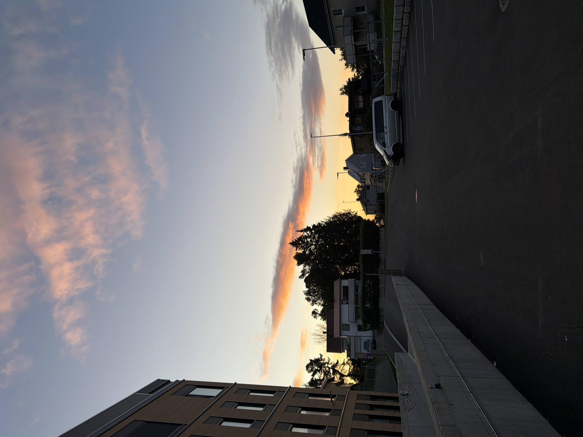 A street scene at dusk in Harstad, Norway. The sky is clear with streaks of pink and orange clouds, indicative of sunset. In the foreground, a dark asphalt parking lot with white parking lines. A silver van is parked in the lot. To the left, a modern building with large windows and beige siding. In the background, several residential houses with pitched roofs and trees. Streetlights are visible. The scene is quiet and peaceful.