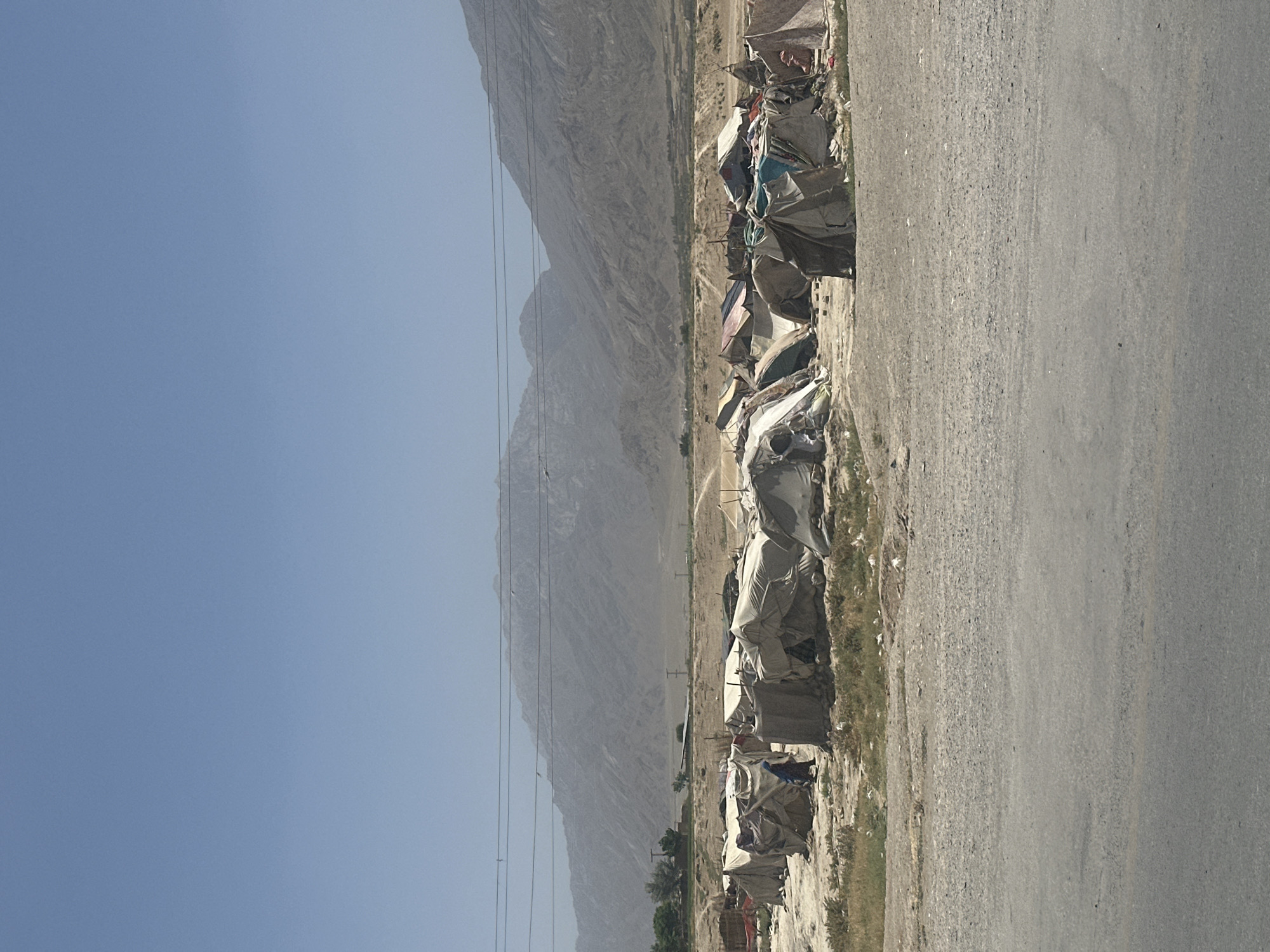 This image depicts an arid landscape in Kuchlak, Pakistan, featuring a roadside settlement against a backdrop of imposing mountains.
In the foreground, a gray asphalt road stretches from the bottom right into the mid-distance. Parallel to the road, on its left side, a strip of dry dirt and sparse, light-colored vegetation marks the immediate ground.
Adjacent to this strip, and extending along the road, is a dense cluster of makeshift shelters. These temporary dwellings are constructed from various scavenged materials such as tarpaulins, plastic sheets, and fabric, draped over frames to form rudimentary homes. They appear weathered and tightly packed, suggesting challenging living conditions.
Beyond the settlement, towering barren mountains dominate the mid-ground, their rocky and eroded slopes stretching across the horizon. These mountains appear hazy due to distance or atmospheric conditions, emphasizing the vastness of the dry landscape. Several thin utility wires or power lines are visible, horizontally traversing the scene above the shelters and in front of the mountain range, supported by barely discernible poles. A faint line of greener vegetation can be observed at the base of the mountains in the far distance, hinting at a valley or different terrain.
The sky above is a vast, clear expanse of light blue, indicating bright daylight, likely midday or early afternoon. The overall atmosphere suggests a hot, dry climate. No people or specific activities are visible within the frame, lending a static, possibly desolate, quality to the scene. There is no visible text.