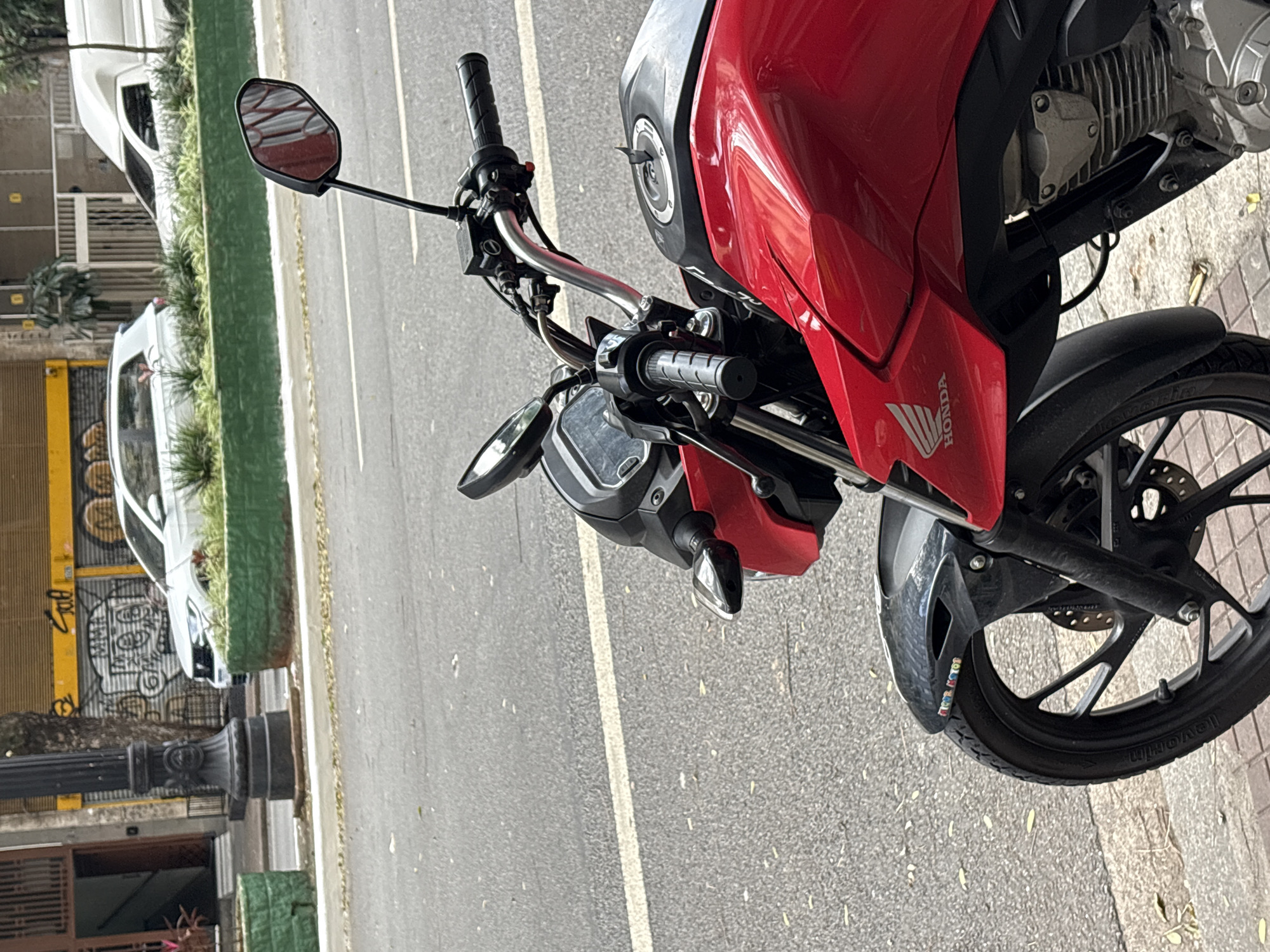 The image shows a red Honda motorcycle parked on a city street. The motorcycle is in the foreground, with its front wheel and part of the engine visible. The handlebars and mirrors are also prominent. In the background, there are cars and buildings, suggesting an urban environment. The street appears to be paved, with a white line marking a lane. Green planters with foliage are visible along the side of the road. The overall impression is a snapshot of a typical day in a Brazilian city.