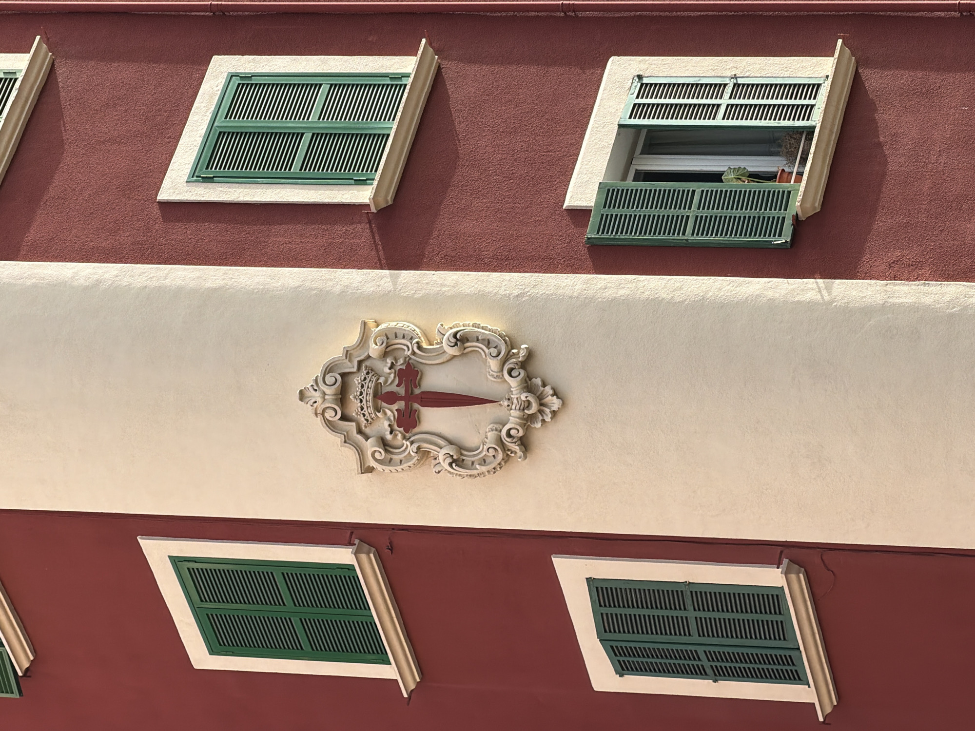The image shows a close-up view of the facade of a building in Palma de Mallorca, Spain. The building has a red exterior wall with a contrasting cream-colored vertical section. There are three windows visible on the red facade, each with green shutters. The top window is partially visible. The middle and bottom windows have their green shutters open, revealing glimpses of the interior.
Centered on the cream-colored section is a decorative, light-colored crest or shield. This crest features an elaborate, ornate design, with scrolling patterns and a crown at the top. Within the crest, a red sword with a cross shape at its hilt is prominently displayed.
The bottom right window has a potted plant on the windowsill, with its leaves visible through the open shutters. The scene suggests a well-maintained architectural feature of a building in an urban setting.