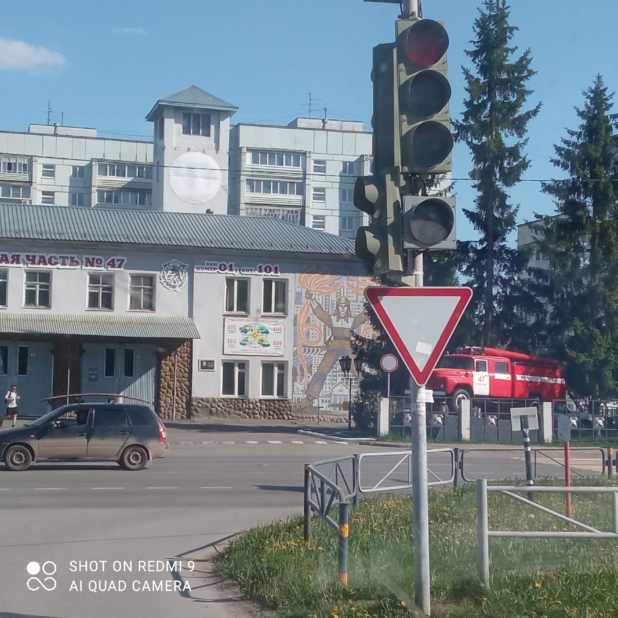 This is an eye-level outdoor shot of a fire station in the city of Chaikovsky, Russia. In front of the fire station, a black station wagon drives to the left. Above and behind the fire station, there is a multi-story building. Next to the street, there is a traffic light that is red. Beside the traffic light is a "yield" sign. Behind the traffic light, a red fire engine is parked at the fire station. On the side of the fire station are several windows and a mosaic image of a firefighter holding hoses. The signage indicates that it is "Fire Station #47" and includes emergency numbers like "101". The setting is urban with green grass in the foreground and trees in the background. The weather appears to be clear and sunny. A person is walking in front of the station.