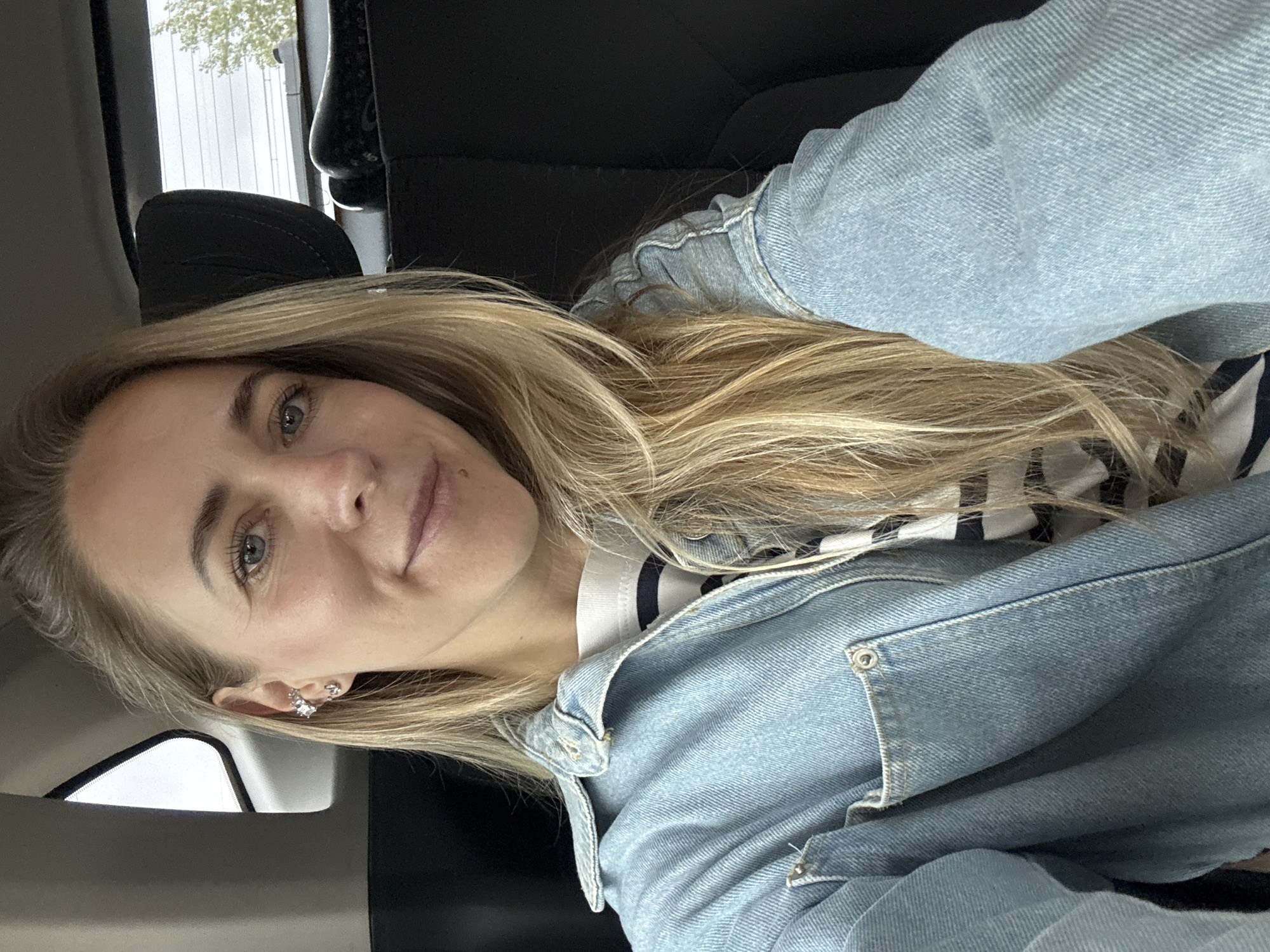 The image shows a close-up, selfie-style shot of a woman inside a vehicle. She has long blonde hair and is wearing a light blue denim jacket over a striped t-shirt. She is also wearing earrings. The background shows the interior of a car, including a headrest and a window with a view of trees and sky. The woman is looking at the camera and smiling slightly. The image does not contain any information that would allow for its location to be identified as Chaikovsky, Russia.