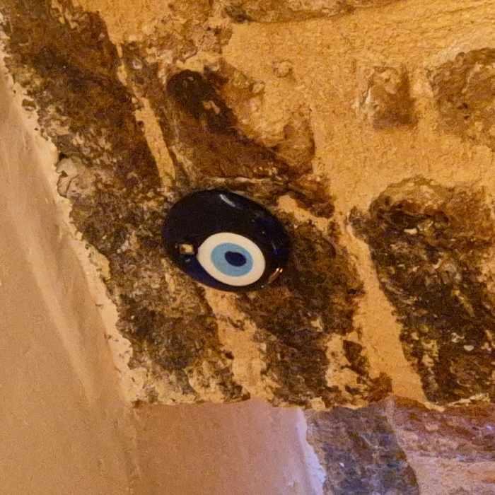 A dark blue, oval-shaped evil eye amulet is attached to a rough, textured surface. The amulet features concentric circles of white and light blue around a central black dot. The background is a mix of beige, brown, and darker areas, possibly stone or brick. The lighting creates shadows and highlights, adding depth to the surface texture. A smooth, light-colored surface is partially visible in the lower left corner.