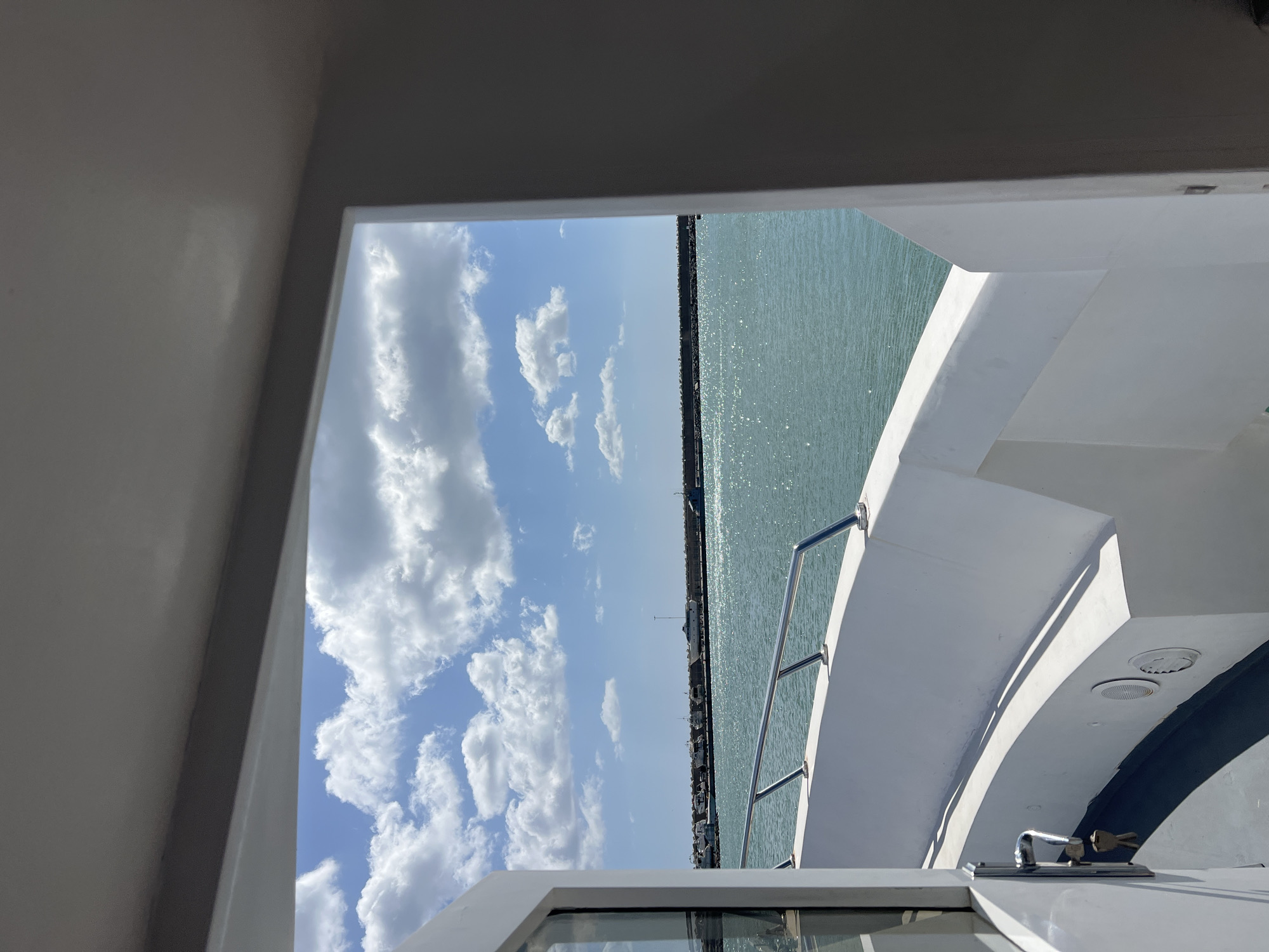 The image captures a view of M'Diq, Morocco, as seen from what appears to be the interior of a boat. The camera is oriented to frame a window or doorway, offering a partial view of the sky, water, and the city's coastline.
The sky is predominantly blue, dotted with white, fluffy clouds. The water glistens with sunlight, and a long pier or breakwater stretches out into the sea, running roughly parallel to the shore. The city's buildings and structures are visible along the coastline, though details are not sharp due to the distance.
The interior elements of the boat are visible, including white walls, railings, and surfaces, indicating a clean, modern design. Some boat hardware, like a latch, is also in view. There are no people visible in the image.
