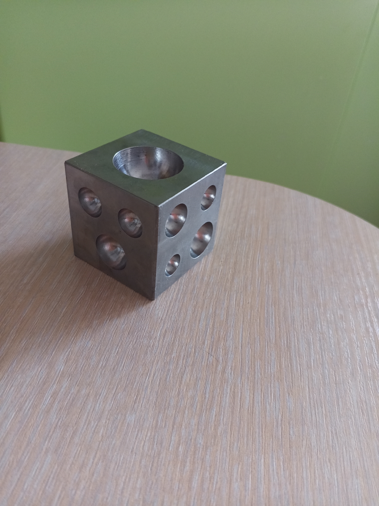 A metal cube with indentations on its faces sits on a wooden table. The cube has a circular indentation on its top face and multiple circular indentations on its visible side faces. The background is a plain green wall. The image is a still life photograph with no people or actions depicted. There is no information in the image to suggest a specific location in Vilyuysk, Russia.
