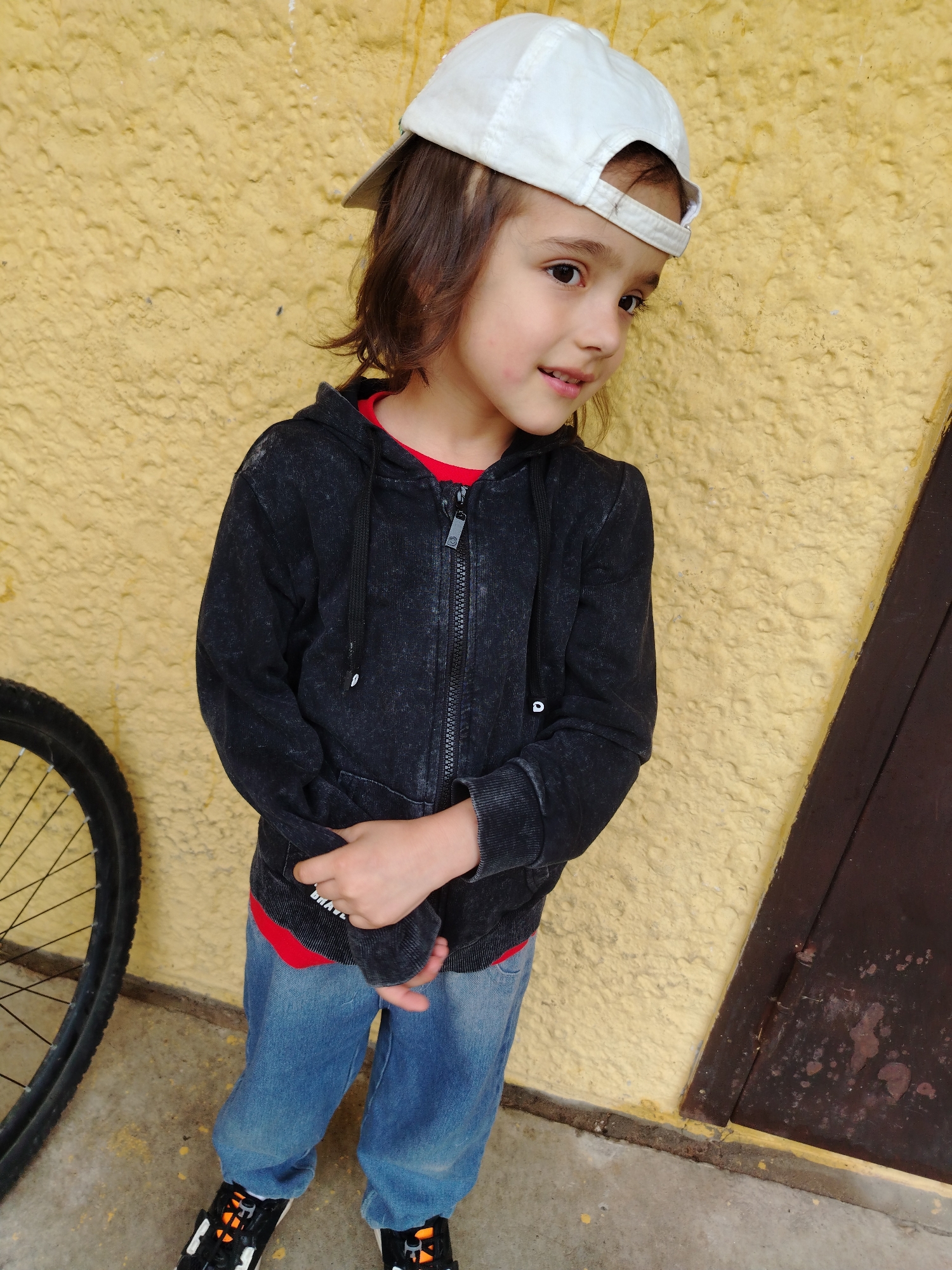 A young person with brown hair, wearing a white baseball cap, a black zip-up hoodie over a red t-shirt, blue jeans, and black and orange sneakers, stands in front of a textured yellow wall. The person is looking to the right with a slight smile. To the left, the wheel of a bicycle is visible. To the right, a dark brown, weathered door is set into the wall. The ground appears to be concrete. The scene suggests an outdoor setting, possibly in a courtyard or alleyway. The lighting indicates it might be daytime. There is no visible text in the image.