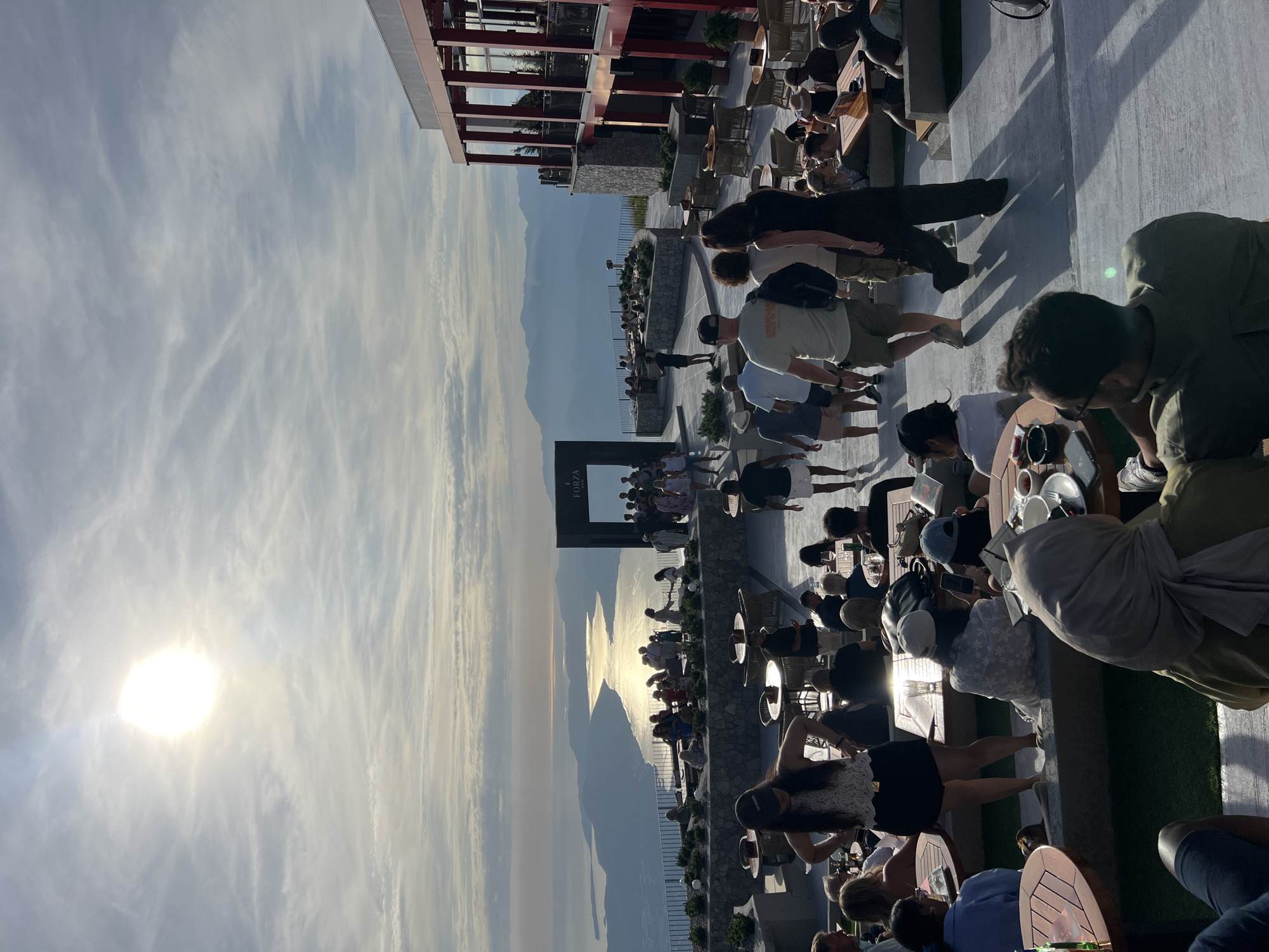 This detailed image depicts a lively outdoor panoramic restaurant or lounge area located in Kuk, Montenegro, during the late afternoon or early evening. The sun, a bright luminous oval, is low in the upper left quadrant of the sky, casting long shadows and a golden reflection across a large body of water that resembles a bay or fjord, bordered by distant mountains.
The foreground and mid-ground are populated by numerous individuals, appearing to be guests and possibly staff, spread across multiple terraced levels. These levels feature a combination of light-colored paved surfaces and sections of artificial green turf. Various round and square tables, made of wood and wicker, are set with matching wicker chairs, where many people are seated. Beverages, including glasses and bottles, are visible on several tables, suggesting dining or socializing activities.
On the right side of the image, a prominent multi-story building with modern architecture features red structural columns and large windows or balconies, indicating an integrated indoor/outdoor facility. In the mid-ground, a large, dark, rectangular archway or gateway, bearing the inscription "FORZA," stands as a notable architectural feature, with a cluster of people gathered around it. Stone planter walls with various small green shrubs and decorative globe lights separate the different terraces.
Numerous people are actively engaged in the scene. Many are seated at tables in groups, interacting, while others are standing or walking across the paved areas. Attire is predominantly casual, suitable for warm weather. Specific observations include: a woman in a white lace top and black shorts standing with hands on hips in the lower left-midground; a man in a light green shirt and baseball cap seen from behind in the lower right-midground, looking right; and a woman wearing a light-colored headscarf seated at a table in the lower right foreground. Several individuals are observed using mobile phones. The low sun creates strong backlighting and prominent, elongated shadows of the people walking, particularly on the paved surfaces.