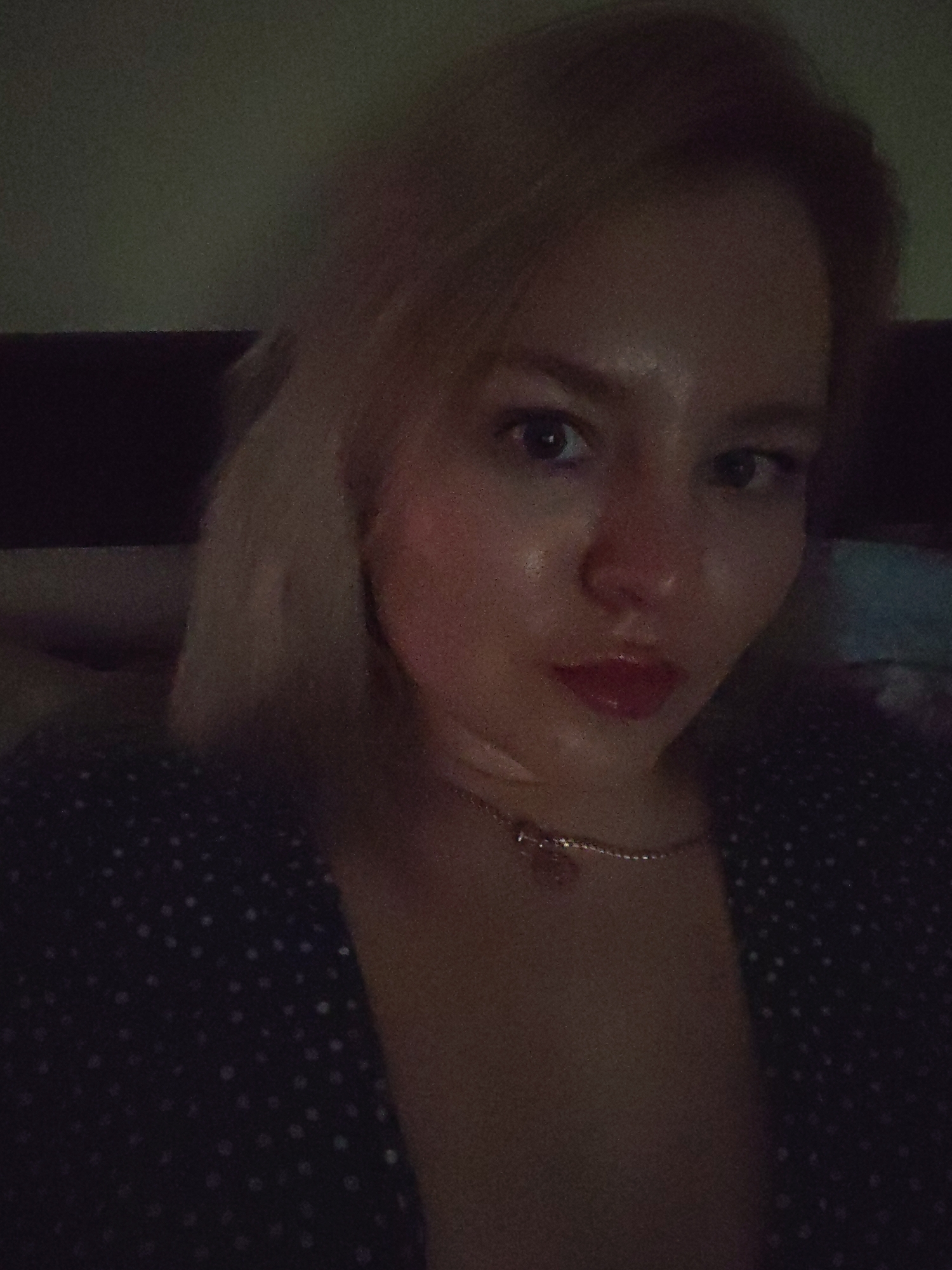 A woman with fair hair and blue eyes is captured in a close-up selfie. She has red lipstick and is wearing a necklace. The background appears to be a headboard and a wall, indicating an indoor setting. She is wearing a dark, polka-dotted top. The image is dimly lit. There is no indication that the image was taken in Izhevsk, Russia.