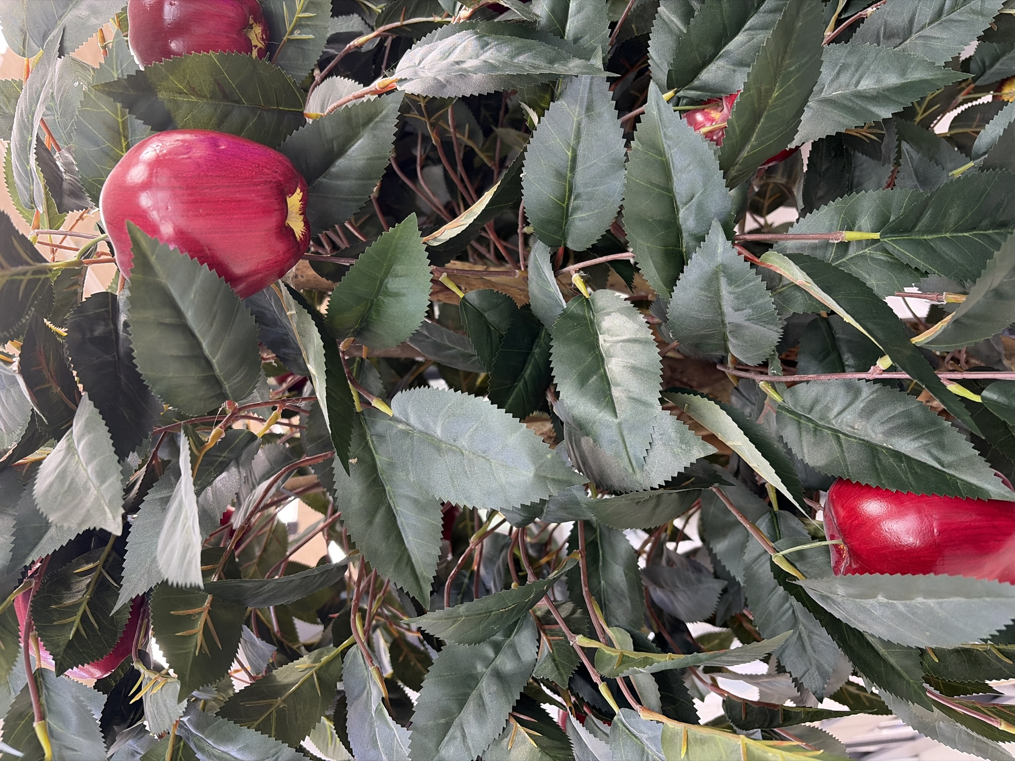 The image displays a detailed, vertical close-up of an artificial apple tree. Numerous dark green, ovate leaves with serrated edges densely cover thin, reddish-brown stems. Interspersed among the foliage are multiple vibrant, deep red artificial apples, characterized by a smooth, glossy texture and a slight yellowish coloration at their bases. Some apples are partially obscured by leaves. No individuals are visible. The background is uniformly blurred, suggesting an indoor environment, possibly a decorative display within a commercial or public space. The specific location within Izhevsk, Russia, cannot be determined from the visual information presented.
