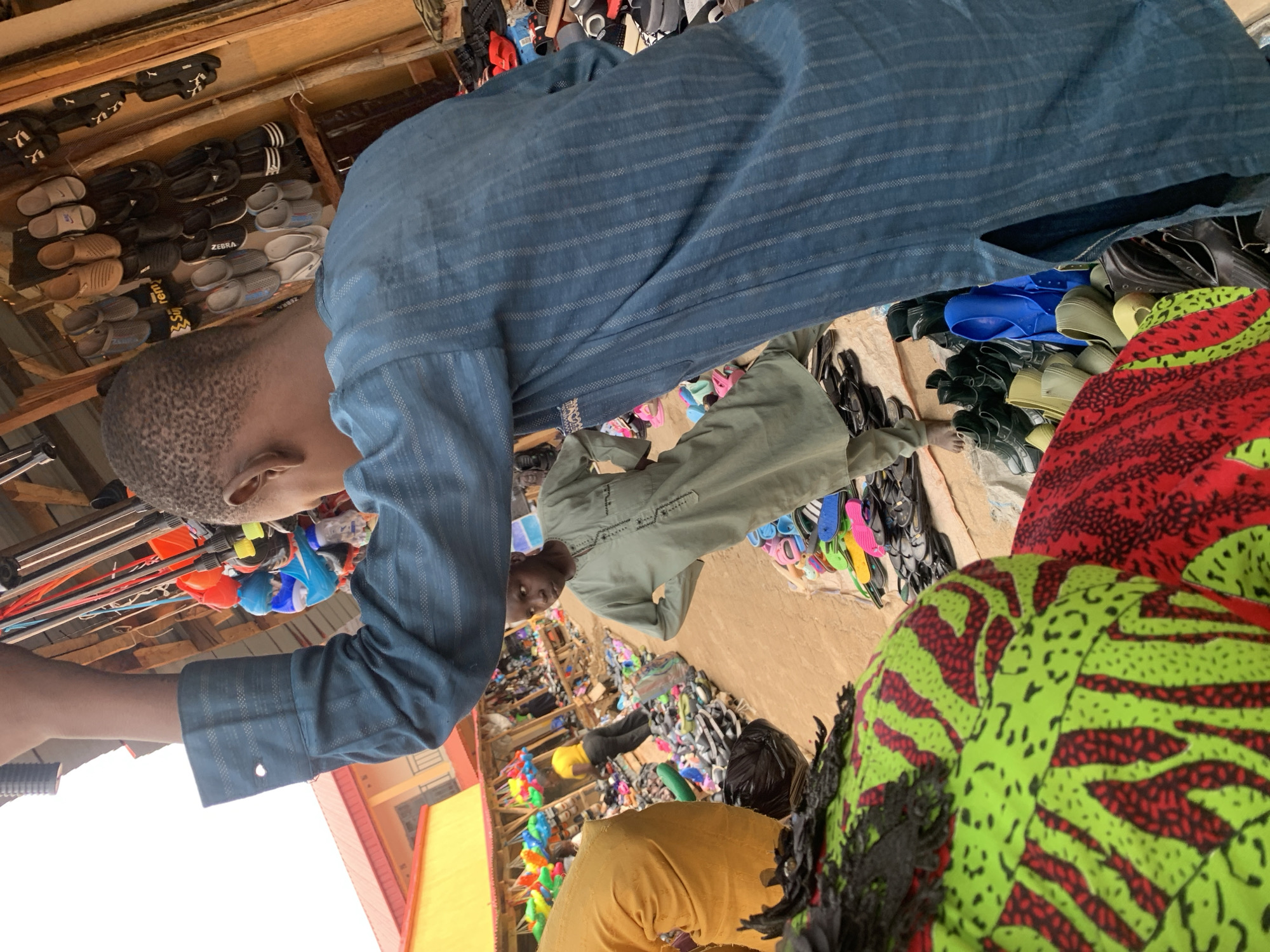 This image shows a street scene in Kafanchan B, Nigeria, with a market selling various goods. People wearing traditional clothing are visible, including individuals in blue, green, and yellow garments. Many sandals and other footwear are displayed for sale. The market setting is characterized by a crowded arrangement of goods.