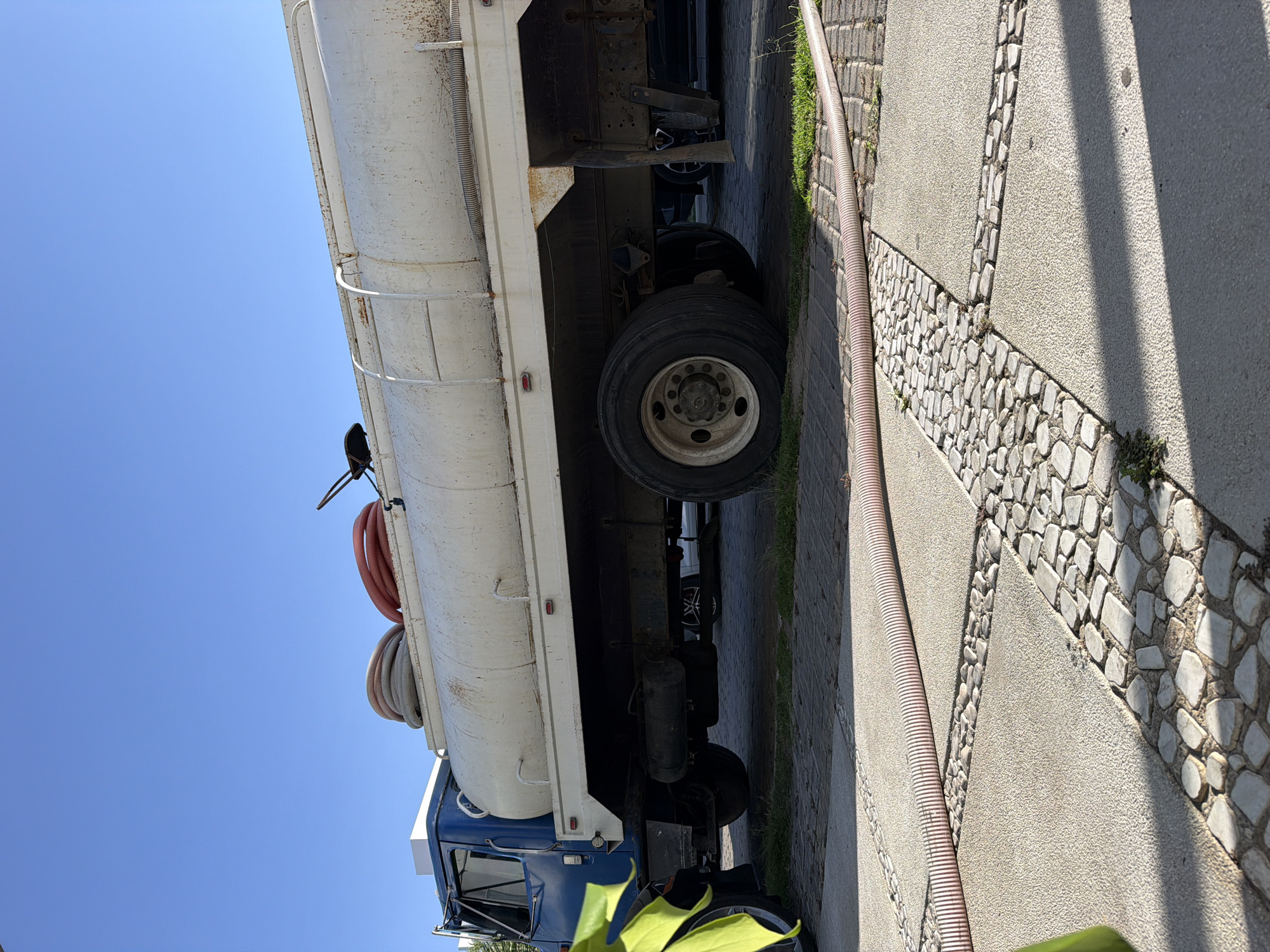 The image, captured in Santa Clara Ocoyucan, Mexico, presents a view of a white tanker truck parked outdoors on a bright, sunny day. The truck features a blue cab and a large, cylindrical white tank, which shows visible signs of wear and rust. Coiled hoses, one reddish-orange and one greyish, are mounted on hooks along the side of the tank. Below the tank, heavy-duty black tires with white hubs are visible, and parts of another vehicle can be seen parked behind it.
On the ground, a long, corrugated hose extends across a distinctively patterned paved area. The pavement consists of light-colored, irregularly shaped stones set into a lighter-toned concrete or aggregate surface, forming decorative strips. Patches of green grass grow along the edges of the pavement. The clear blue sky dominates the upper portion of the frame, indicating bright, clear weather conditions. Strong shadows are cast on the ground, suggesting it is daytime. A yellow or lime-green high-visibility garment is partially visible in the lower foreground, implying the presence of a person nearby, although no individual is fully seen. No discernible text is visible in the image.