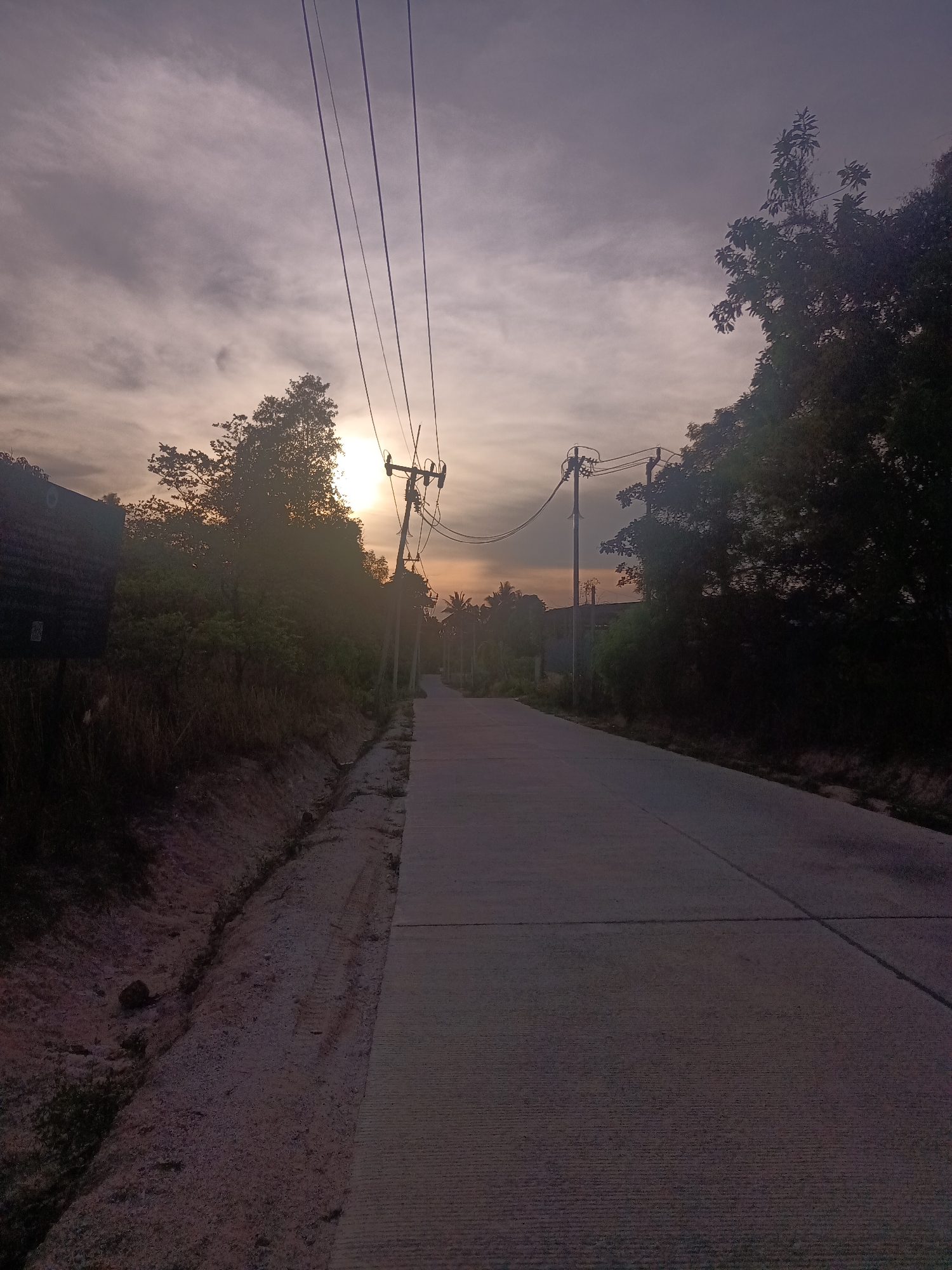 The image depicts a road in Moo 4, Thailand, at either dawn or dusk. The sky is overcast with the sun appearing behind trees in the distance. The road is concrete and stretches into the distance. Power lines run along the side of the road, supported by poles. Vegetation, including trees and shrubs, lines both sides of the road. A drainage ditch runs along the side of the road on the left. A dark rectangular sign is visible on the left. The overall lighting is subdued.