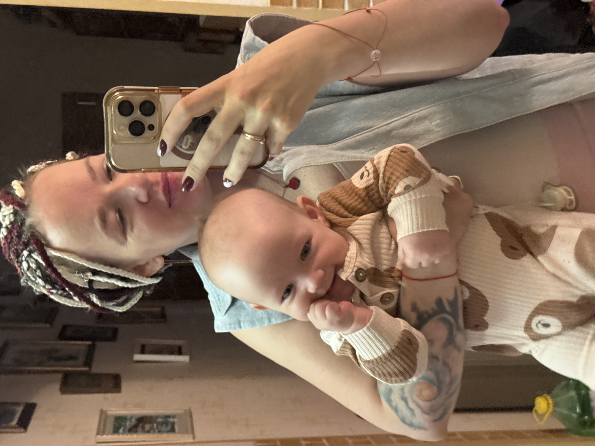A mirror selfie captures a woman holding a baby in an indoor setting, possibly a home in Moscow, Russia. The image is rotated 90 degrees clockwise.
The woman, seen in reflection, has light skin, braided hair in shades of white, brown, and reddish-purple, and dark nail polish with white accents on her fingernails. She is wearing a light-colored shirt, possibly a denim or chambray style, over a lighter undershirt. A gold-toned ring adorns her left ring finger, and a delicate red string bracelet with a clear bead is on her right wrist. A tattoo with a swirling, abstract design is visible on her lower right arm. She is holding a gold-colored smartphone with three camera lenses to take the picture, and her expression suggests a soft smile or contentment.
The baby, held close by the woman, has light skin and is wearing a light-colored, ribbed romper with brown stripes and cartoonish bear or sloth patterns. Buttons are visible down the front of the romper. The baby is awake, looking towards the camera, and has one hand near its mouth, possibly teething or exploring.
In the background reflection, a wall with several framed pictures of varying sizes is visible. Below the baby, a light-colored surface is seen, with a pacifier resting on it. A green-capped bottle is partially visible in the bottom right corner of the reflection. The overall scene conveys a warm, affectionate moment between the parent and child. No discernible text is visible.