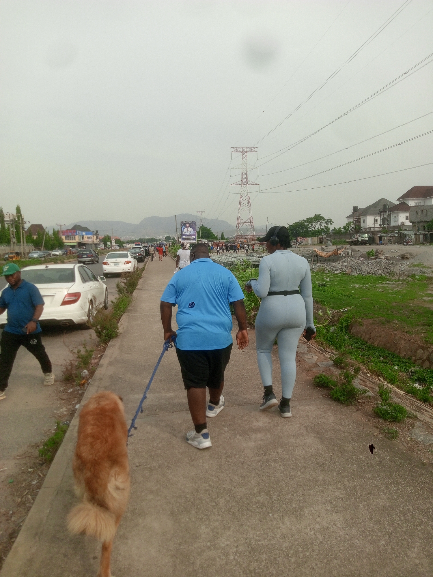 The image depicts a scene in Dushepe, Nigeria. A man in a blue t-shirt and black shorts is walking a golden-colored dog on a leash along a concrete walkway. To his right, a woman wearing a light blue jumpsuit and headphones walks beside him. They are moving away from the camera. The walkway is bordered by a grassy area on one side and a road with parked cars on the other. Other people can be seen walking in the distance. Overhead, there are power lines supported by a red and white transmission tower. A mountain is visible in the background.