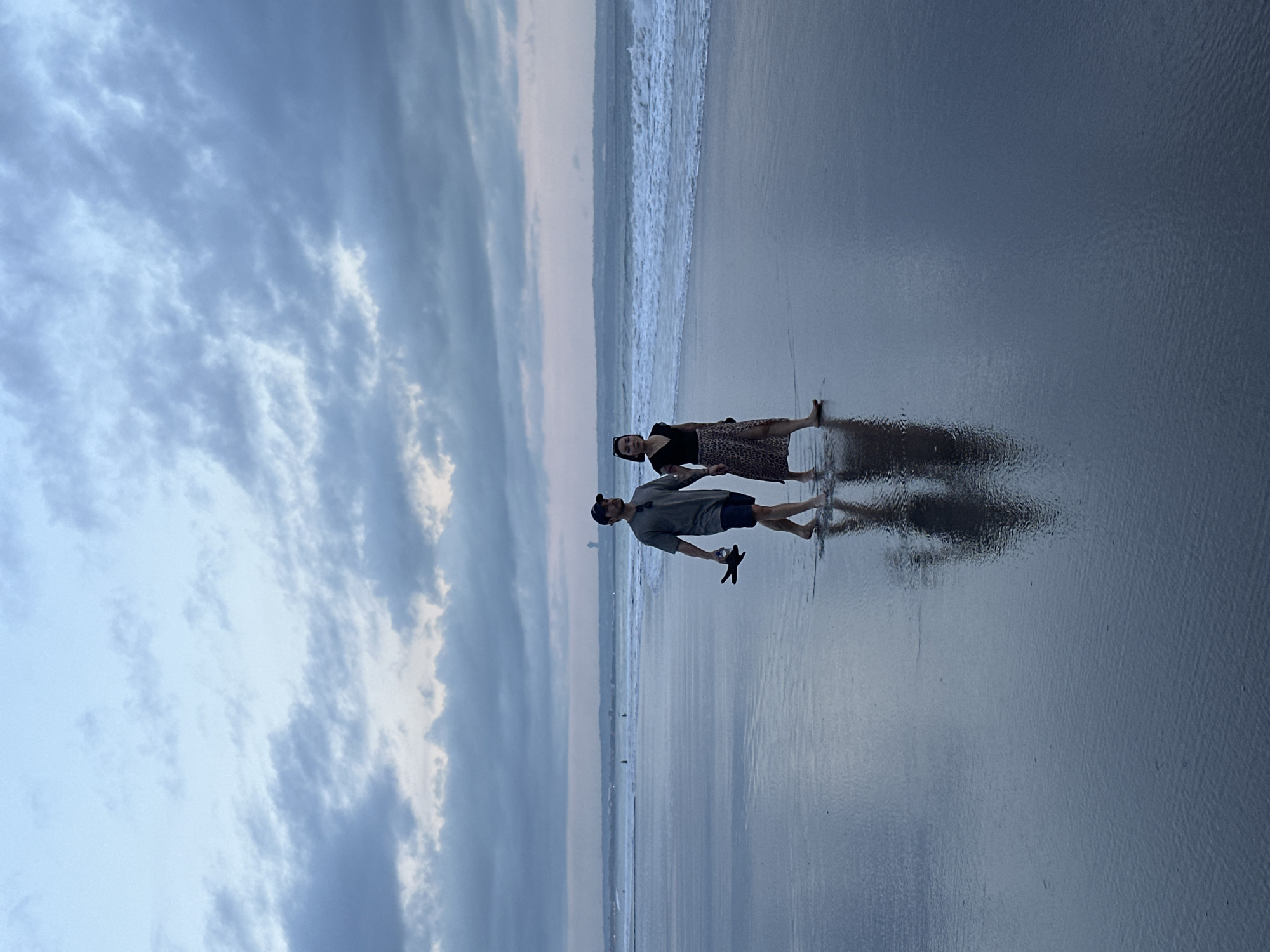 On a reflective beach in North Kuta, Indonesia, a man and a woman stand near the water's edge under a cloudy sky. The woman is wearing a dark top and a patterned skirt. The man wears a light-colored shirt, dark shorts, and a cap and holds a black object in his hand. The sky is covered with gray and white clouds, and the ocean is visible behind the couple.