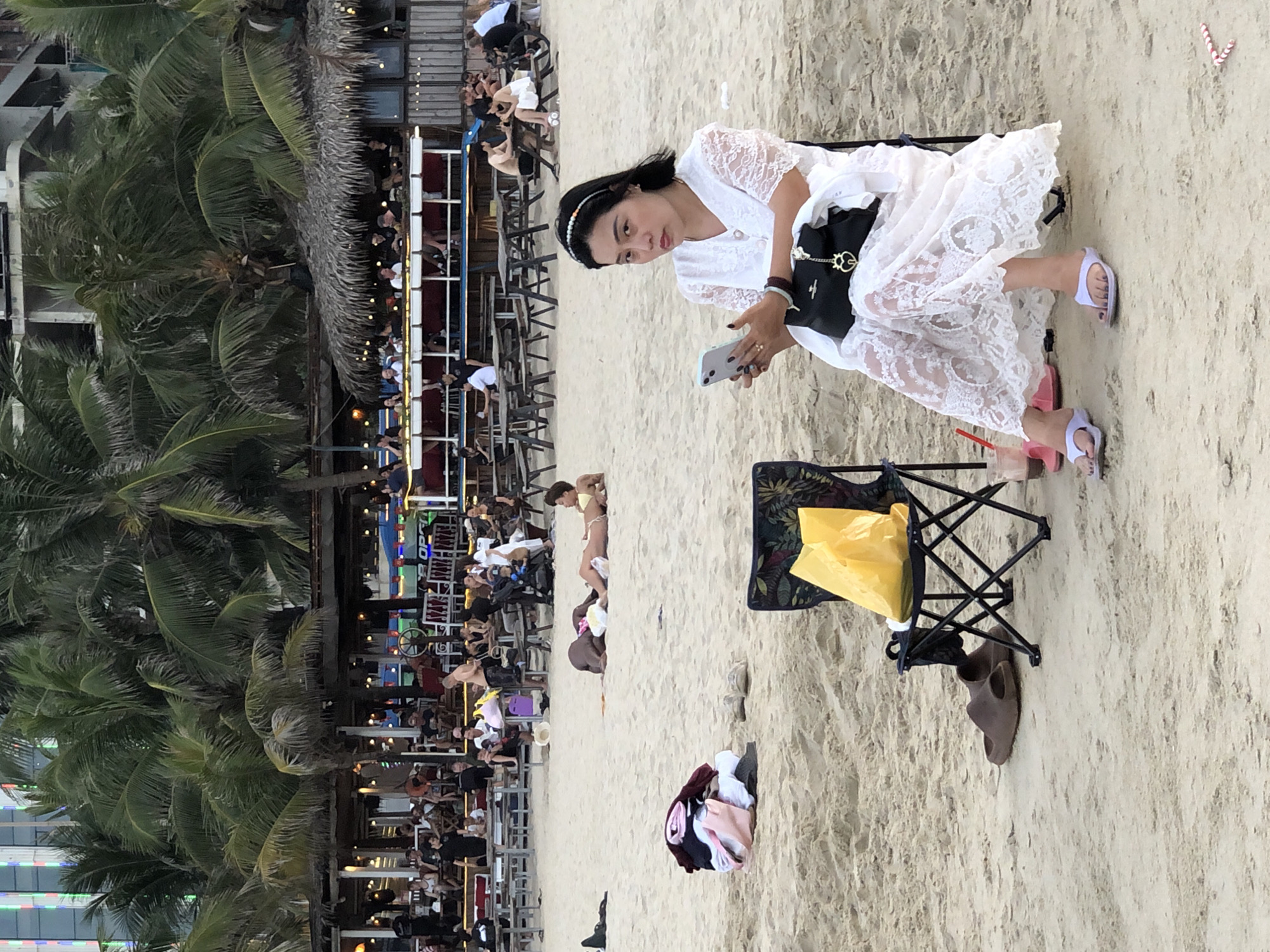 This image captures a scene at My Khe Beach in Da Nang, Vietnam. In the foreground, a woman is seated on a beach chair, dressed in a white lace dress, and holding a smartphone. Beside her is a black handbag. Her feet are adorned with light purple flip-flops. A yellow plastic bag is placed on the chair. Further in the background, several people are lounging on the sand. One person is lying on their stomach wearing a yellow bikini. In the middle ground, a pile of folded clothing rests on the sand. Behind them, a bustling beachfront establishment with thatched roofs and wooden seating areas is visible, with numerous patrons enjoying the ambiance. Palm trees frame the scene, and buildings can be seen in the distance. The overall environment suggests a warm, tropical day at the beach.
