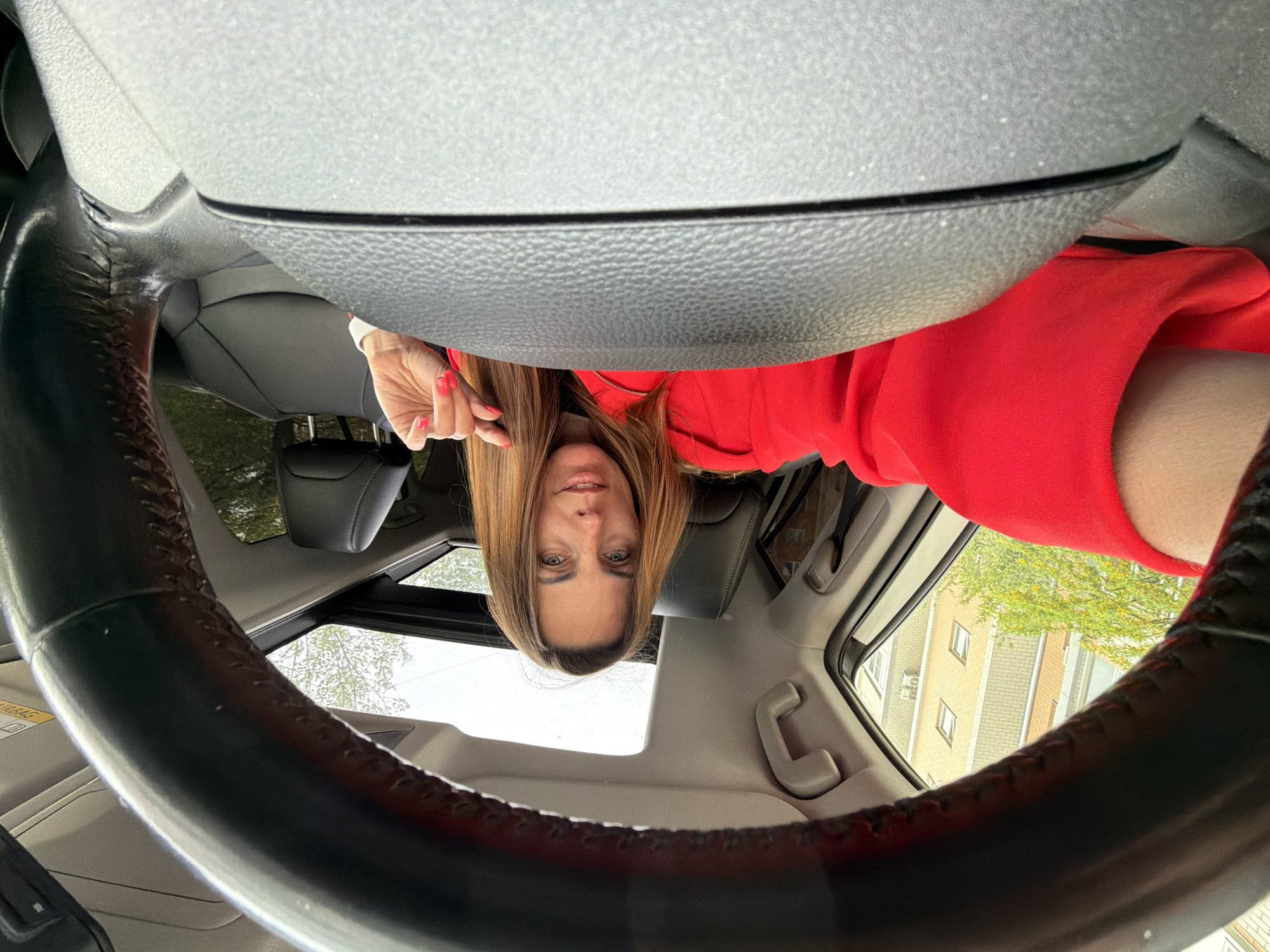 A woman with long brown hair, wearing a red top, is in a car. She is behind the steering wheel, and the image is taken from a low angle, looking up. Through the car's sunroof, a building is visible, potentially located in the city of Birobidzhan, Russia. The car's interior includes gray seats. The woman is holding up her hand.