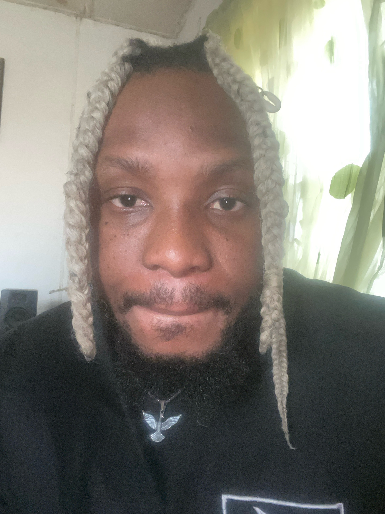 The image is a close-up, selfie-style portrait of a Black person with pale blonde braids and a full beard. The subject is wearing a black hoodie with a silver bird pendant. The background appears to be an indoor setting with a white wall and a green curtain or sheer fabric. There is a speaker visible to the left of the subject. The lighting is bright, possibly from a window, and casts a soft glow on the subject's face. There are no explicit geographical markers in the image to definitively place it in Jos, Nigeria. However, the overall aesthetic and the presence of the speaker could be consistent with a domestic setting in a city like Jos.