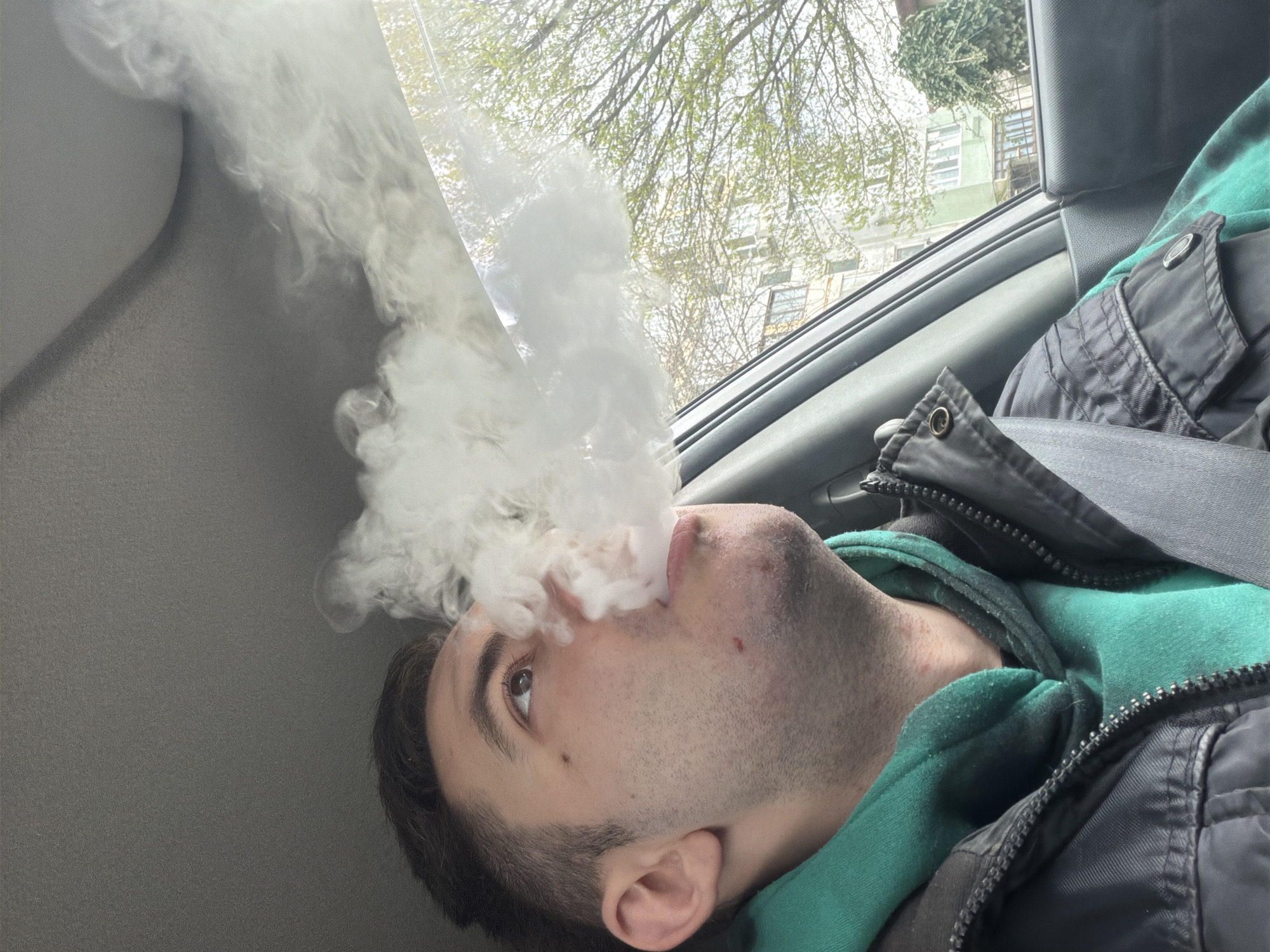 A young man, likely in his early twenties, exhales a large cloud of white vapor. He is seated in the passenger seat of a car, wearing a green hoodie under a black jacket. The car's window shows a glimpse of buildings and trees, potentially located in Varna, Bulgaria. The buildings seen are light colored, with white or light green paint. The trees are sparsely leafed, suggesting early spring. The interior of the car is grey. The image appears to be taken from a low angle and rotated to the side.