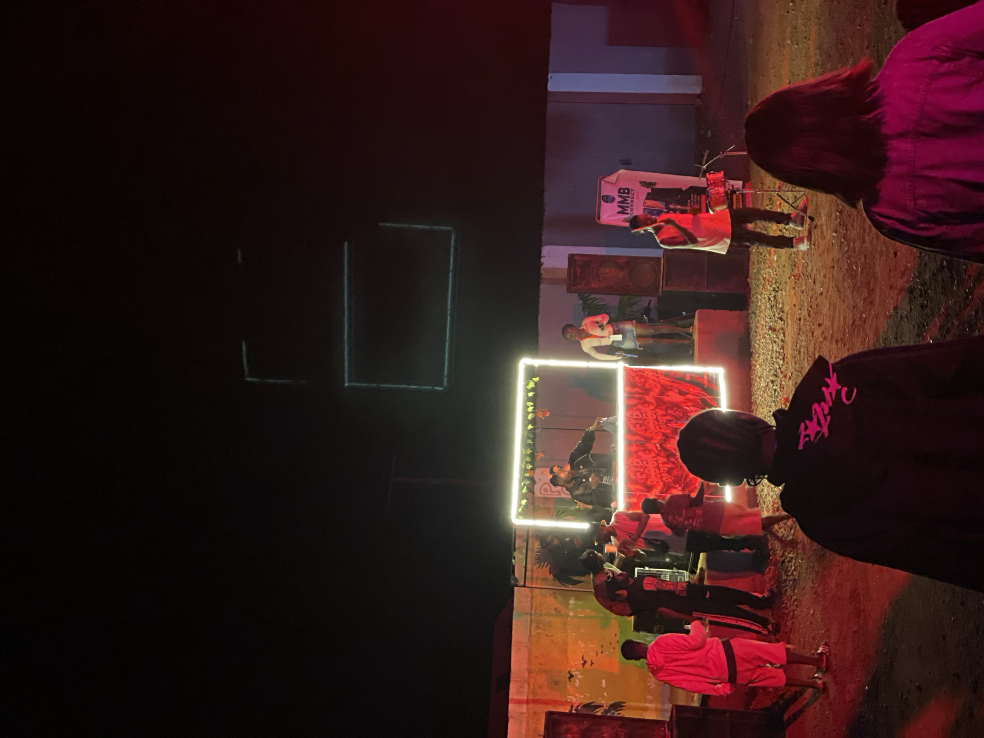An outdoor nighttime event is taking place in Durumi, Nigeria, with a focus on a stage area featuring a DJ booth and performers. The scene is illuminated by neon lights and red stage lighting, casting a vibrant glow on the surroundings.
The DJ booth is centrally located, enclosed by a neon light frame and decorated with red lighting and a patterned facade. Two DJs are visible behind the booth, actively engaged with their equipment. To the left of the DJ booth, a female performer in a white top and denim skirt stands holding a microphone, appearing to address the audience.
In front of the stage, a small audience is gathered. A group of individuals in the foreground are partially visible, with their backs to the camera, observing the performance. To the left of the stage, a person in a pink hoodie and white shorts stands facing away from the camera. Further left, a group of three individuals are interacting near some sound equipment.
To the right of the stage, a man in a white tank top and dark pants is standing with a microphone, appearing to be part of the performance. Behind him, a banner with the text "MMB CONNECT" is displayed. Several large speaker boxes are positioned around the stage area.
The environment appears to be a somewhat unpaved outdoor space, with dirt and gravel on the ground. A textured wall with graffiti or artwork is visible behind the stage area. The overall atmosphere suggests a lively music event or concert.
