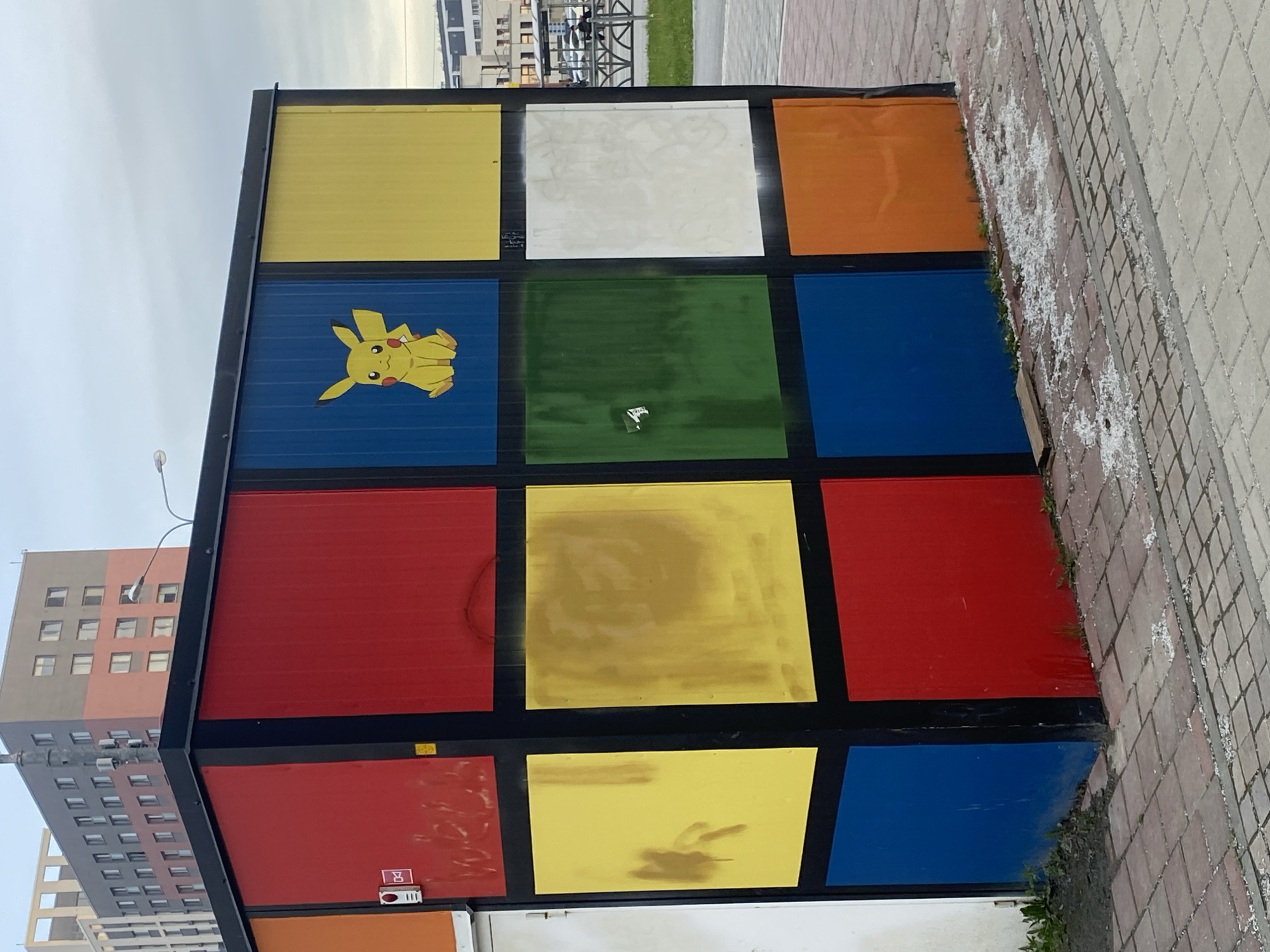 The image shows a multi-colored cube-shaped structure resembling a Rubik's cube. Each face of the cube is a different color, including yellow, white, orange, blue, red, and green. There is a Pikachu character depicted on one of the blue faces. The structure appears to be located in Yekaterinburg, Russia, as suggested by information from image analysis. The cube sits on or near a sidewalk composed of interlocking paving stones. A building is visible in the background, its architecture suggesting a contemporary urban setting. The sky is overcast, contributing to diffused lighting. The environment appears to be an outdoor public space.