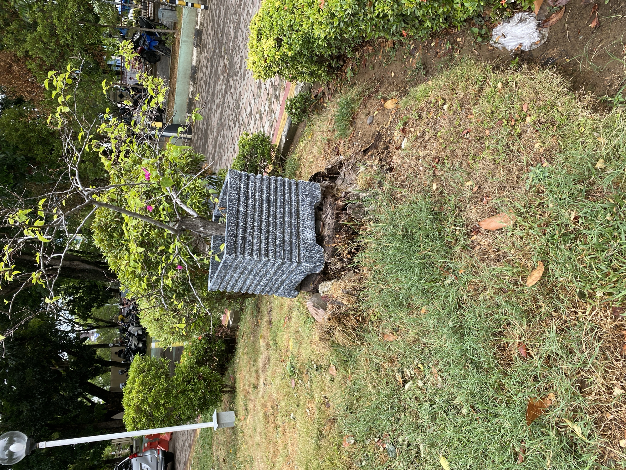 In Banda Aceh, Indonesia, a decorative potted tree with a gray, textured planter rests on a tree stump in a grassy area. The planter is square with horizontal ridges. The tree has sparse, light-green leaves and a few small pink flowers. To the left of the planter, a white lamppost with a round, globe-shaped light is visible, partially obscured by a neatly trimmed green bush. Behind the planter, several motorcycles are parked in what appears to be a paved area with a decorative border. In the background, more trees and foliage are present, suggesting an outdoor public space like a park or courtyard. The ground in the foreground is covered with grass, some of which is dry and brown, interspersed with fallen leaves. To the right, another green bush is visible, and a piece of white plastic trash is lying on the ground near a narrow dirt path.