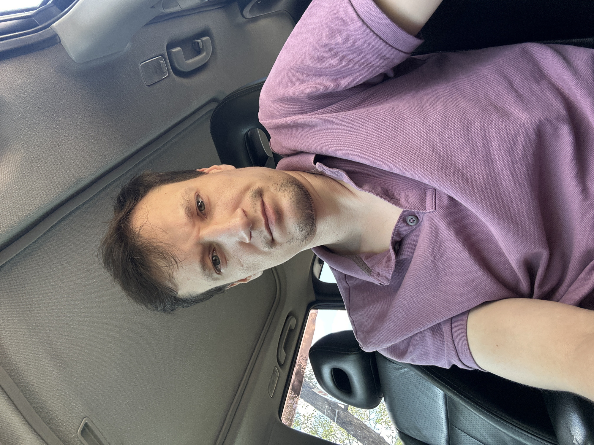 A man is sitting in the driver's seat of a car, looking towards the camera. He is wearing a short-sleeved purple polo shirt. The interior of the car is visible, including the headrest of the passenger seat, the car's ceiling with a sunroof, and a portion of the dashboard. Outside the car window, trees and sunlight are visible. The image appears to have been taken during the daytime. The location is the city of Makiv, Ukraine.