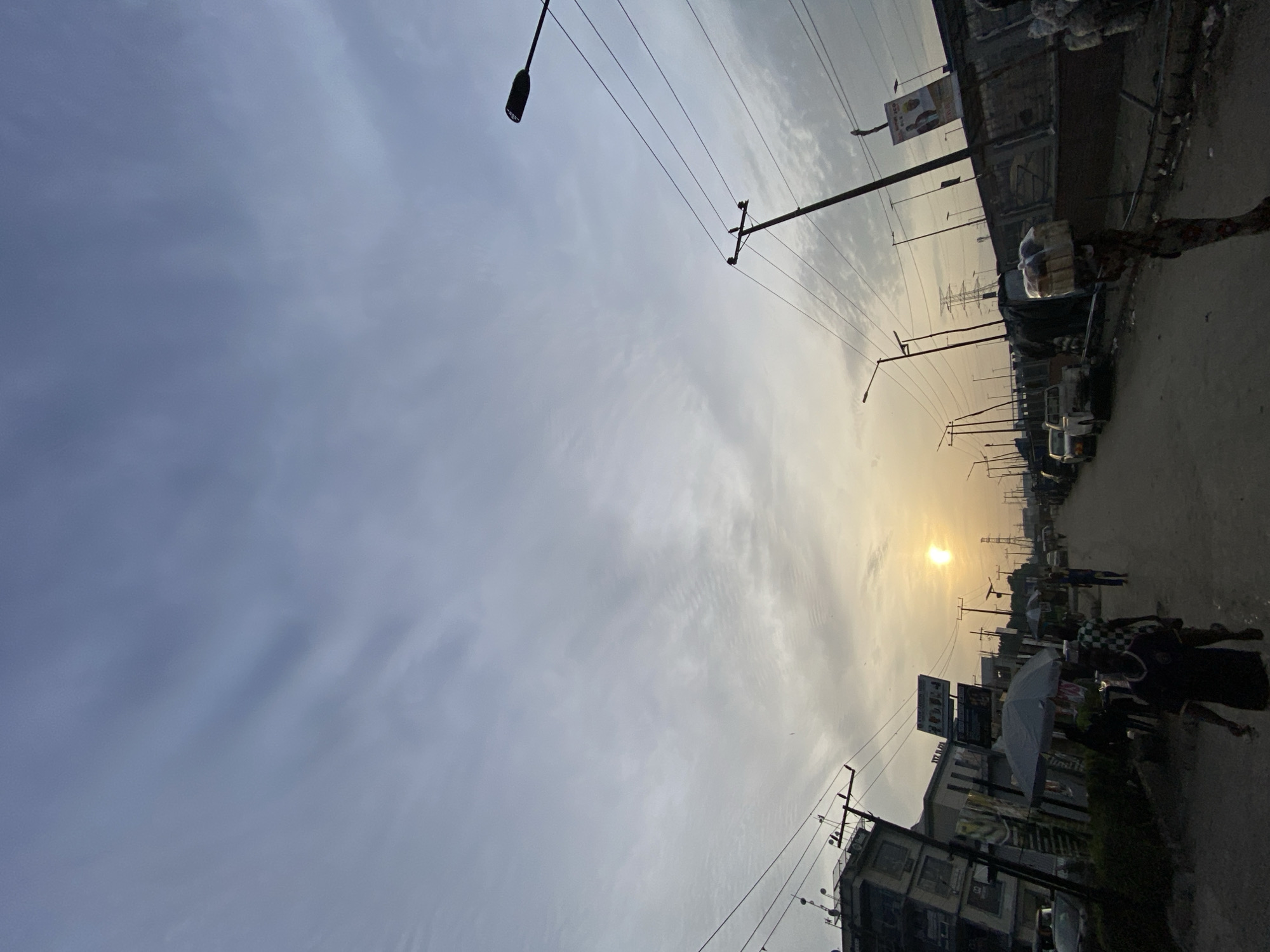 This media file depicts an outdoor street scene in Ado, Nigeria, at either sunrise or sunset, indicated by the low, golden sun prominent on the right side of the frame. The sky is largely overcast with thin, streaky clouds, but the weather appears clear.
The scene features a street lined with buildings, numerous utility poles, and an intricate network of overhead power lines. Several people are visible engaging in daily activities along the street. In the lower right corner, a person walks holding an open, light-colored umbrella. Further down the street, additional indistinct figures can be seen, alongside parked vehicles, including what appears to be a white truck.
Various commercial signs are affixed to buildings and poles. On a building in the foreground, a large black billboard-style sign is visible with white text, including "TREND" and "HOTEL," and another sign below it indicates a "Lounge." Another banner-like sign on a utility pole displays graphics suggesting food or snacks. The street surface appears to be a mix of paved and unpaved sections. The overall impression is one of a bustling urban or semi-urban area during the golden hour.