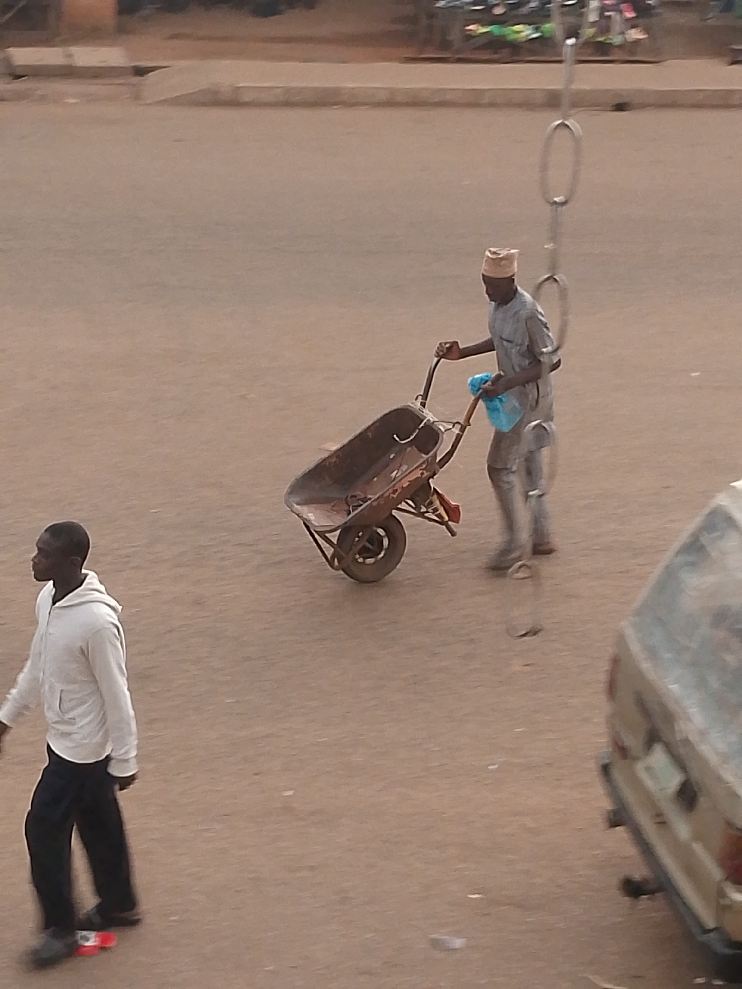 In Bukuru, Nigeria, an overhead shot captures two individuals on a dusty, outdoor surface that appears to be a road or marketplace. On the left, a person wearing a white hooded sweatshirt and dark pants walks away from the camera, their feet shod in dark shoes. Notably, the person is standing on a red and white object that resembles a skateboard.
Further to the right, another individual, dressed in a light-colored, patterned tunic and a cap, is pushing a traditional metal wheelbarrow. The wheelbarrow appears to be loaded or used for carrying items. This person is holding a bright blue plastic bag in their left hand. In the background, a portion of a light-colored vehicle is visible on the right side of the frame. The overall environment is urban, with a muted color palette dominated by browns and grays, suggesting an outdoor setting in Nigeria.