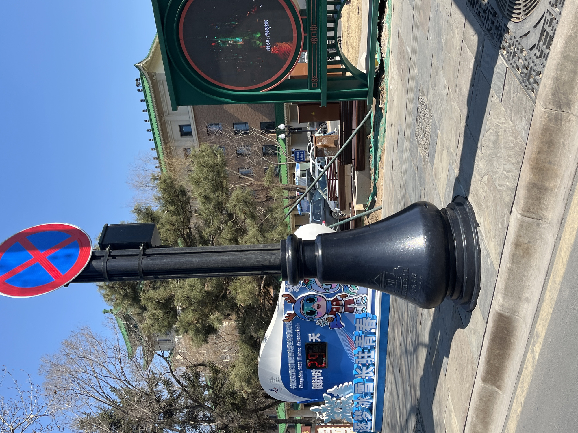 The image captures a street scene in Changchun City, China, under a clear blue sky. Prominently featured is a black metal lamp post, affixed with a "no stopping" traffic sign. Adjacent to the lamp post is a blue and white sign promoting the Changchun 2027 Winter Universiade. The sign features a cartoon mascot and indicates a countdown of 1 day. The temperature is 12 degrees Celsius. The building in the background, adorned with a traditional green-tiled roof, suggests a blend of modern and historical architectural styles. The ground is paved with grey stone. A large circular screen with a green frame is placed on a tall green metal structure. A large tree and a few parked cars are visible behind the sign.