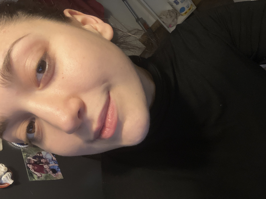 A close-up selfie captures a person with fair skin, brown eyes, and arched eyebrows. Light reflects on their face, accentuating features like the nose and lips, which are colored with a pink tint. They wear a black shirt. In the background, there are miscellaneous objects including a photo of a group of people.