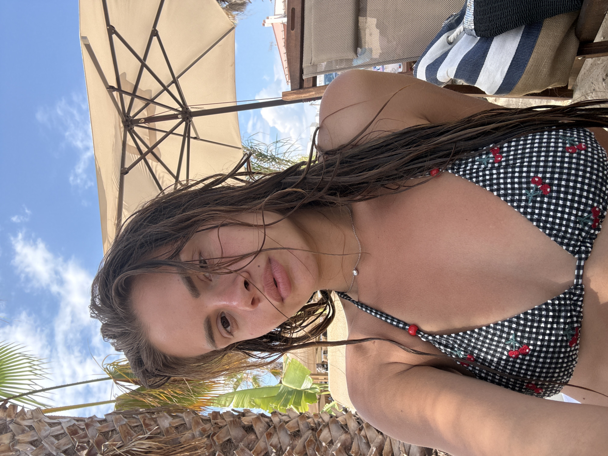 A young woman with wet, dark hair is taking a selfie on what appears to be Ofrynio Beach in Greece. She is wearing a black and white checkered bikini with a cherry pattern. The background shows a blue sky with white clouds, a palm tree, a large beige beach umbrella, and a chair. Sunlight is casting shadows on her face and body.