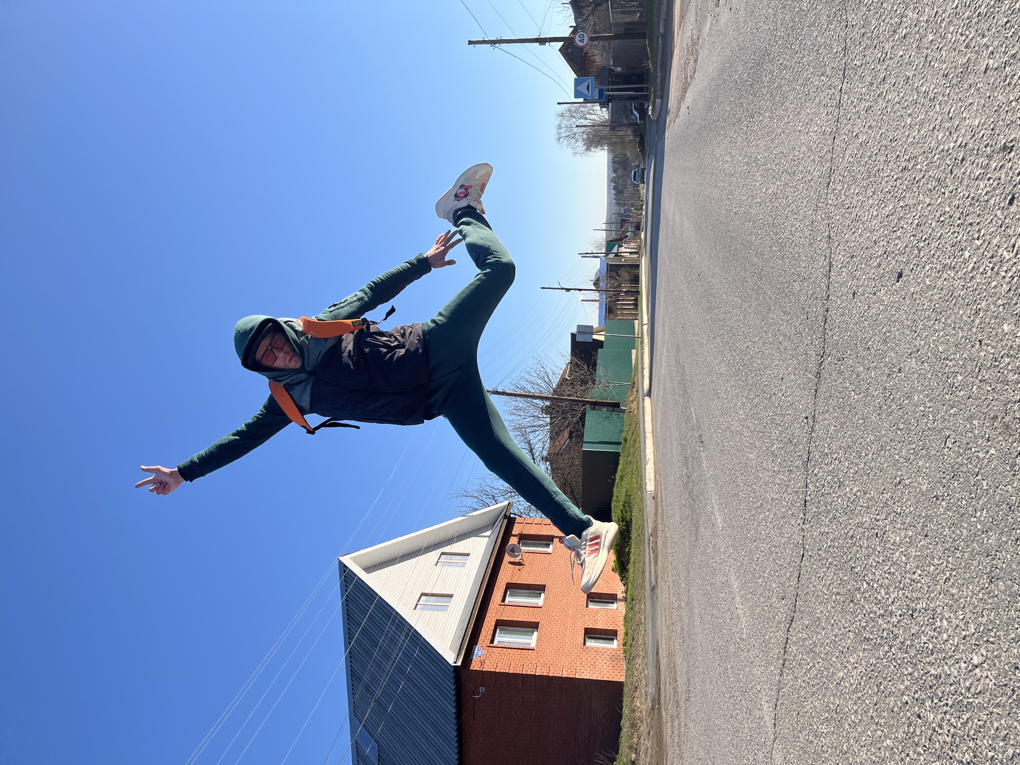 This media file captures a dynamic scene on a clear, sunny day in what appears to be a residential or semi-rural area of Chaikovsky, Russia. The image is taken from a low angle, looking upwards.
The central subject is a person, likely male, mid-jump or mid-action, suspended in the air. They are wearing a dark green hooded jacket or hoodie, dark green athletic pants, and white sneakers with distinctive red stripes or accents. Sunglasses cover their eyes, and an orange-strapped backpack or harness is visible over their jacket. Their arms are outstretched, one pointing forward, the other slightly bent, while one leg is bent high in the air and the other is also bent, contributing to a striking, energetic pose.
The background features a bright, cloudless blue sky dominating the upper portion of the frame. Below the person, a grey asphalt road stretches into the distance on the right, showing a visible crack down its center. To the left, a two-story house with red brick walls and a distinctive gabled roof, partially covered in white and grey siding, is visible. Further down the road on the right, more houses, utility poles, and overhead power lines can be seen. A circular speed limit sign displaying "40" (presumably 40 km/h) is clearly visible on a pole, along with other unreadable signs. The sparse trees in the background appear to be largely bare, suggesting spring or autumn.
The overall impression is one of movement, joy, or athleticism, captured under brilliant sunlight.