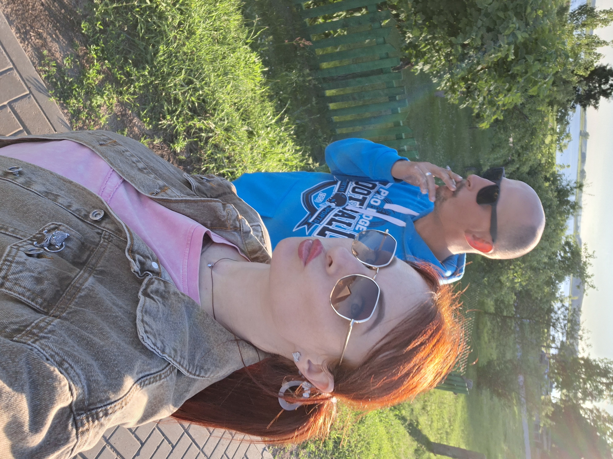Two individuals are in the foreground and mid-ground of the image. The person in the foreground, a female, is positioned on the right side, facing the camera, and has red hair pulled back into a ponytail. She is wearing a light pink shirt, a denim jacket, sunglasses with a hexagonal frame, and a necklace with a small pendant. She is making a kissing gesture. The person in the mid-ground, a male, is on the left side, facing away from the camera and slightly to the side. He is bald, wearing a blue hoodie with "PRO HOGE FOOTBALL" printed on it, and black sunglasses. He is smoking a cigarette. The background shows a green wooden fence, lush green grass, trees, and a body of water in the distance, suggesting an outdoor, possibly park-like or recreational area. The lighting indicates it is daytime, possibly late afternoon given the warm glow. The cobblestone pathway in the foreground suggests a developed area within a park or recreational space. The overall setting evokes a casual, outdoor leisure activity. The location is specified as Smaliavičy, Belarus, which could be a small city or town known for its natural surroundings and recreational areas.