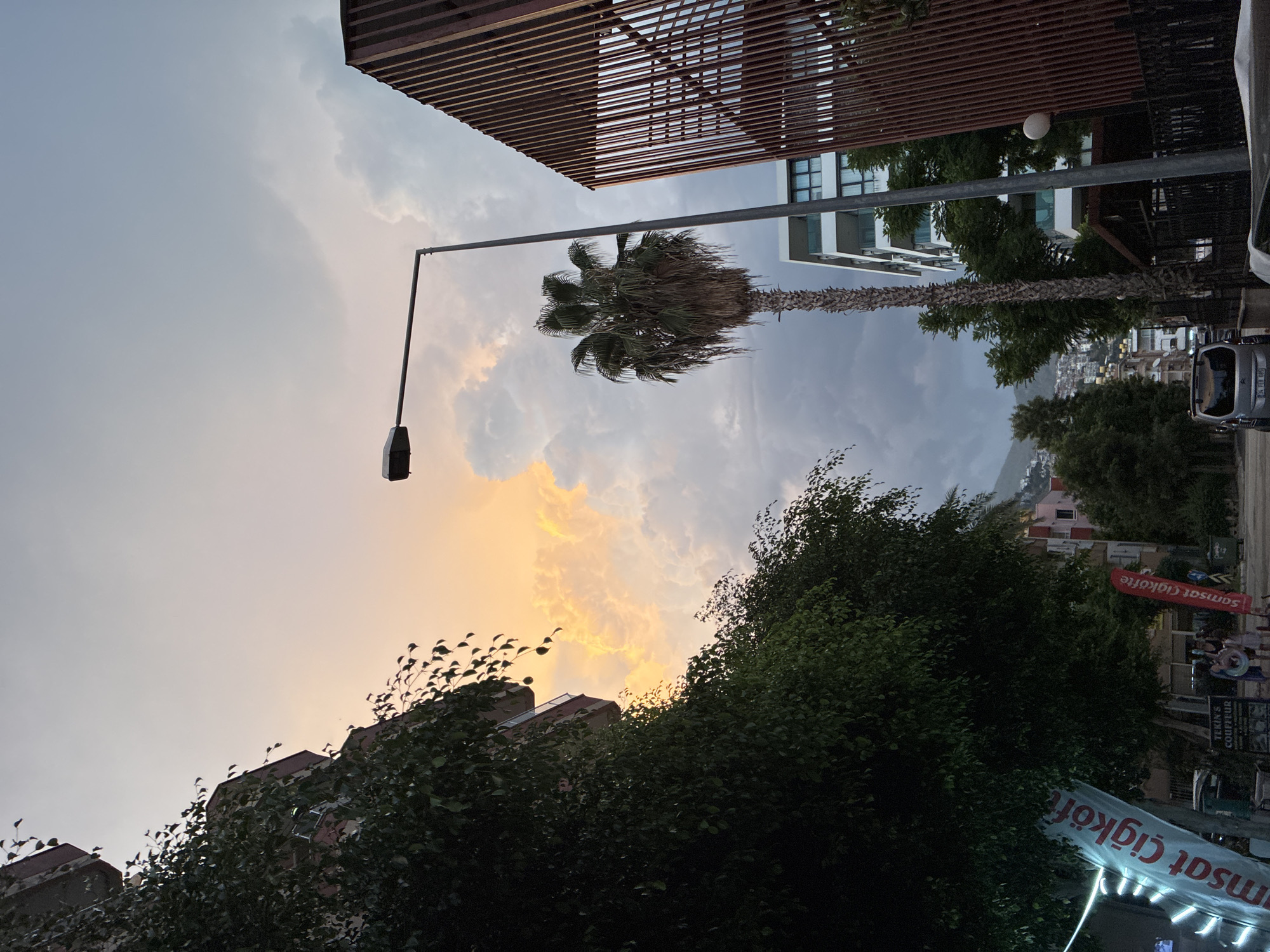 This is a street-level, low-angle, full-shot photograph taken in Alanya, Turkey, during what appears to be sunset or dusk. The sky is filled with dramatic clouds, illuminated by the setting sun in a vibrant orange and yellow hue, contrasted with darker, grey clouds on the right.
On the left side of the frame, a multi-story building is partially visible, heavily covered in dark green foliage. A street sign or banner, predominantly blue with white text, is attached to the building, reading "samsat Çiğköfte".
In the center of the image, a tall street lamp with a single light fixture stands prominently. Beside it is a mature palm tree with a slender trunk and a full crown of fronds.
To the right, a modern building with a distinctive facade composed of vertical, reddish-brown slats rises. Further back, a white, multi-story building with balconies and glass windows is visible, surrounded by trees.
In the background, a hillside covered with buildings and greenery slopes upwards, leading towards a mountainous landscape under the darkening sky.
In the foreground, on the street, a grey minivan is parked. Two individuals are walking on the sidewalk near a storefront that appears to be a hairdresser with a sign reading "TEKIN'S COIFFEUR". Another red banner, similar to the blue one, also reads "samsat Çiğköfte". The street is paved and appears to be lined with shops and trees.