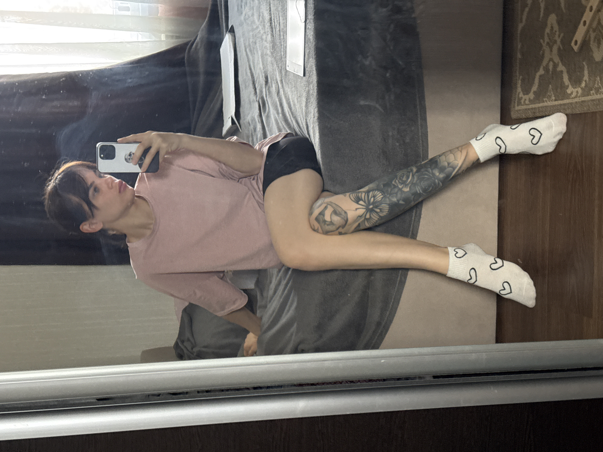 A young person is taking a selfie in a mirror. The individual is lying on a grey bed or couch. They are wearing a pink t-shirt, black shorts, and white socks with black hearts. The person has tattoos on their leg. The background includes a window with sheer curtains and a rug on the floor. The image appears to be indoors. There is no indication that this image was taken in Rostov-on-Don, Russia.