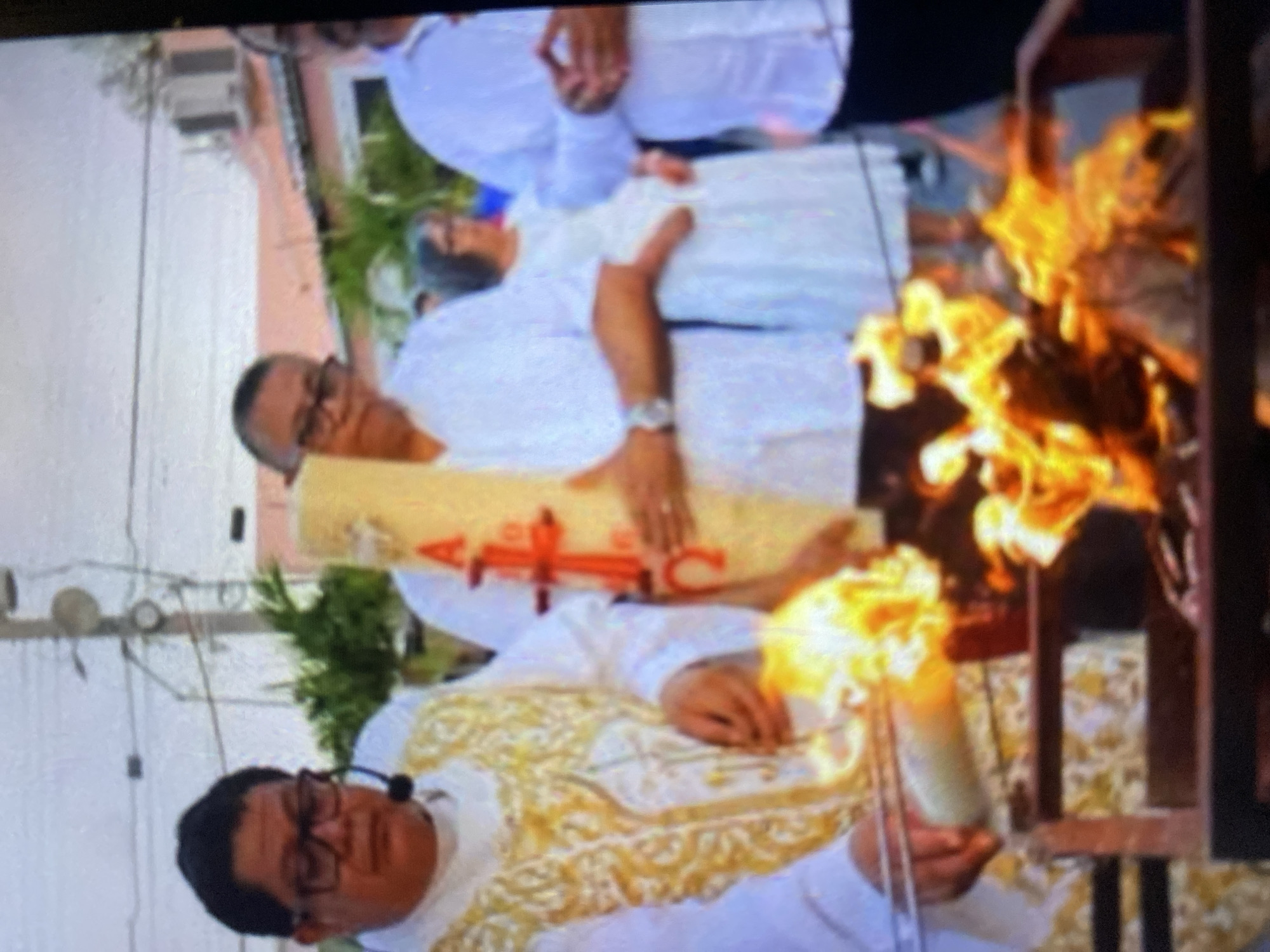 The image depicts a scene in Tinaquillo, Venezuela. A Catholic priest in a white robe with gold trim stands holding a lit candle and tongs next to a fire pit containing burning materials. A deceased person dressed in white lies in state on a yellow-draped bier, attended by mourners dressed in white. The surrounding environment includes a street with utility poles and overhead wires, a building with an air conditioning unit, and some greenery.