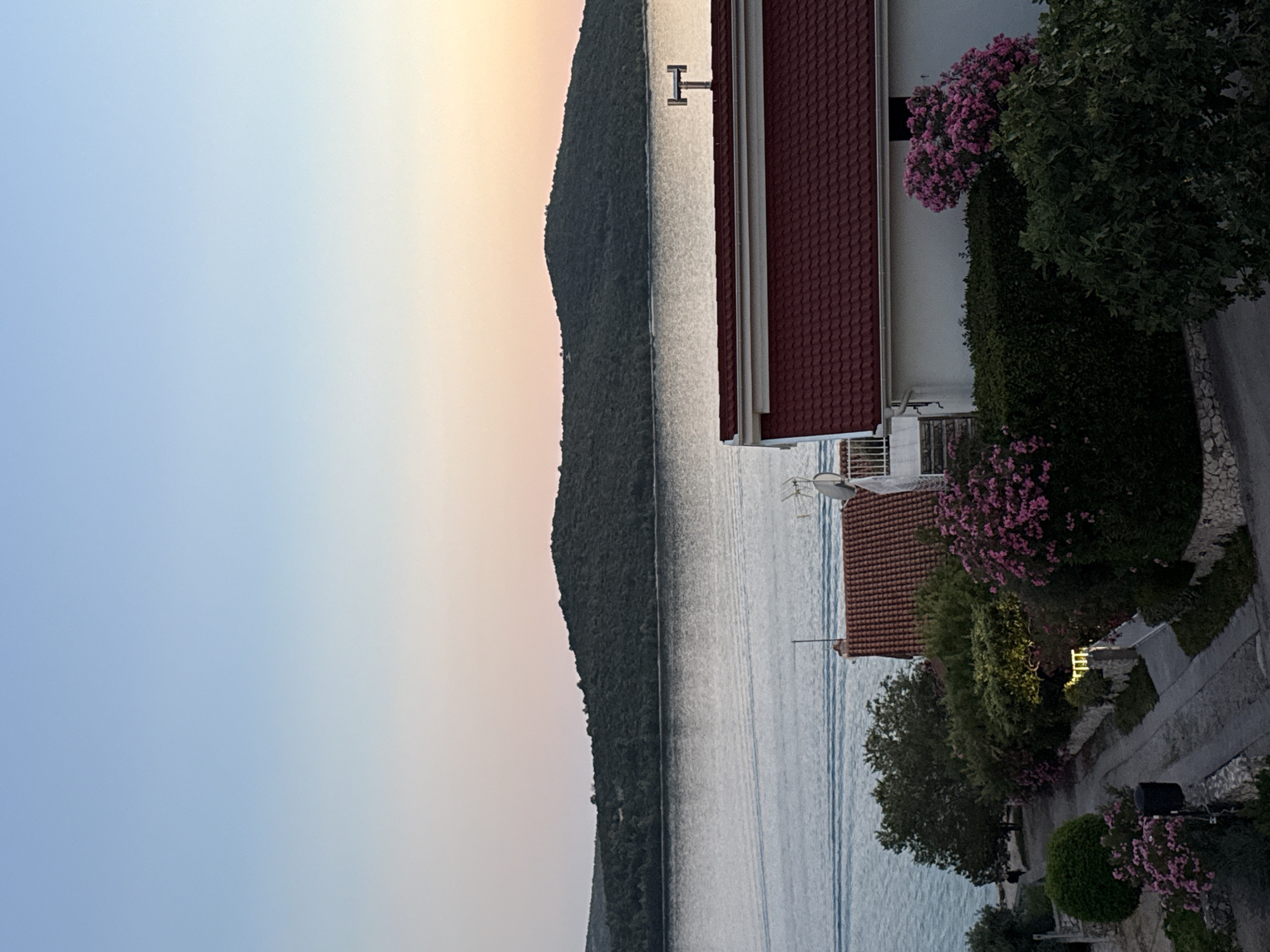 The image depicts a serene coastal landscape at dusk, likely in or near Grad Šibenik, Croatia. In the foreground, a cluster of houses with distinctive red tiled roofs is nestled amongst lush greenery and vibrant pink flowering bushes. A narrow, paved pathway winds through the scene, bordered by stone walls and manicured hedges. Beyond the houses, a tranquil body of water, likely the Adriatic Sea, reflects the soft hues of the setting sun. In the distance, a densely forested island rises from the water, its silhouette a dark green against the pale, gradient sky that transitions from a clear blue at the top to soft oranges and pinks near the horizon. The overall impression is one of peaceful domesticity harmoniously integrated with the natural beauty of the Croatian coastline.