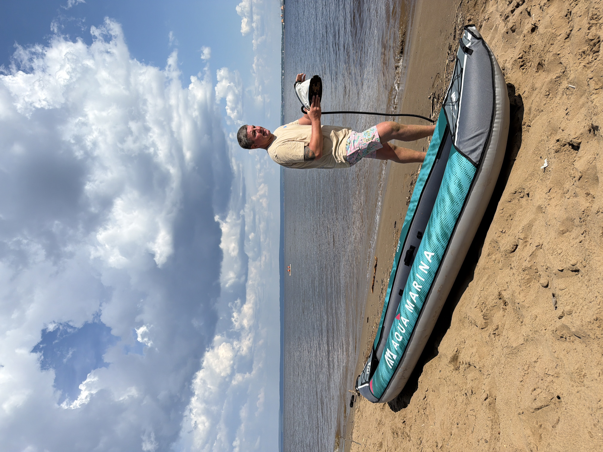A man stands on a sandy beach near the water, holding a white and black object with a black cord attached. He is wearing a beige t-shirt and patterned shorts. In front of him on the sand is an inflatable kayak with the brand name "Aqua Marina" visible. The kayak is teal and gray. The water is dark and calm, with a few distant boats visible. The sky is blue with large, fluffy clouds. The location is identified as Noviy, Russia.