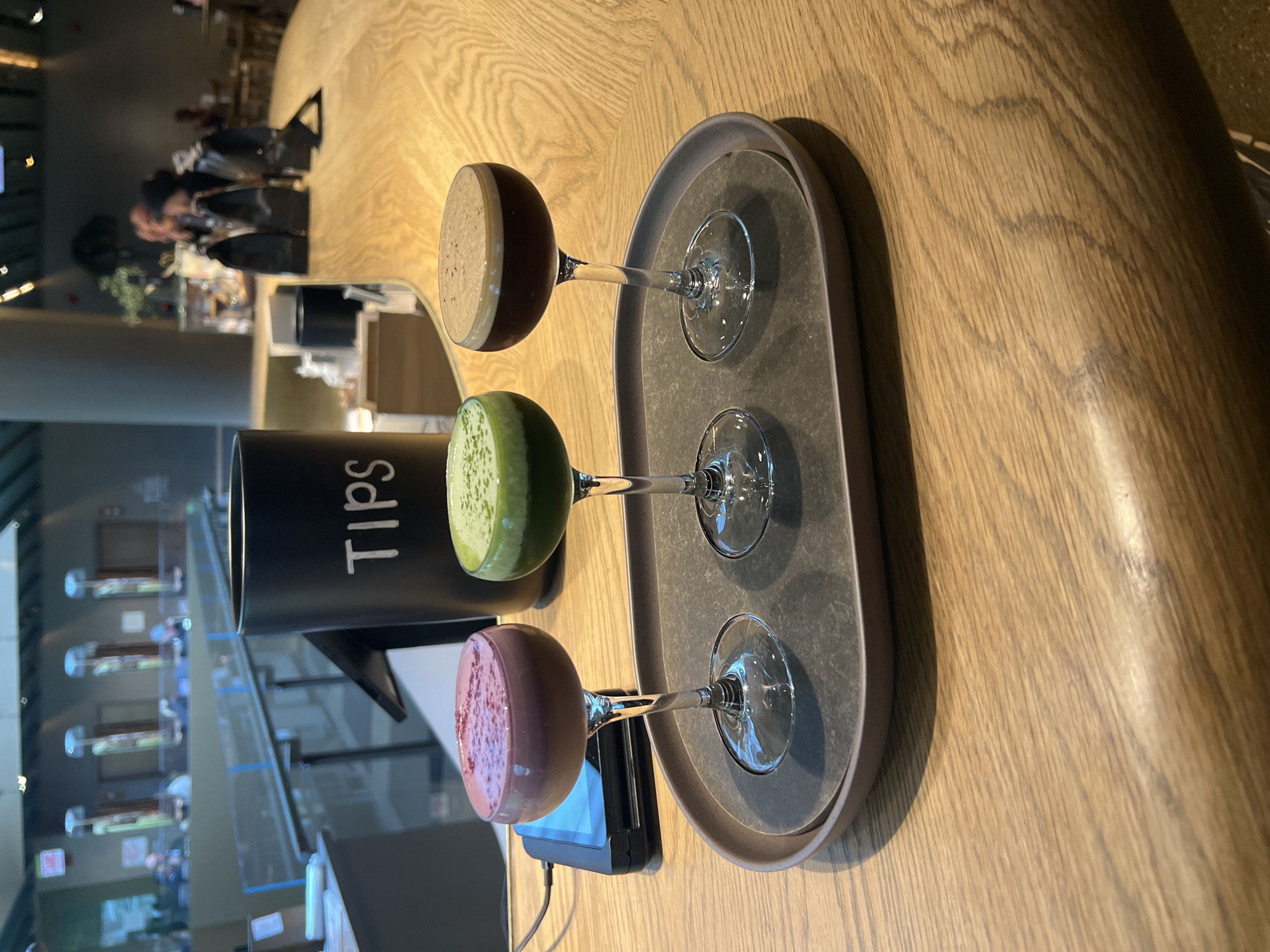 The image depicts three stemmed glasses filled with different colored liquids - purple, green, and brown – arranged on a gray oval tray. A black container labeled "Tips" is positioned beside the glasses. In the background, there is a blurred view of people sitting at tables and the interior of what appears to be a restaurant or bar. The location appears to be the city of Chicago, United States of America, due to the architectural style of the building visible through the glass window and some visible signage. A payment terminal is visible next to the glass of pink liquid. The tray sits on a light brown wooden surface, possibly a countertop or table.
