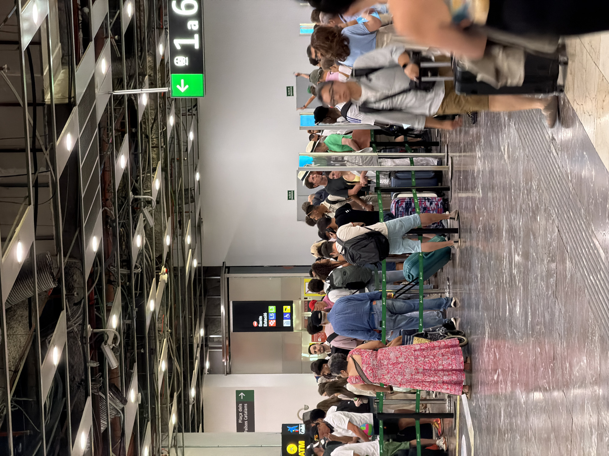 The image captures a busy scene inside a train station or airport in Barcelona, Spain. A large group of people are standing in queues, indicating waiting for boarding or a service. The environment features a polished floor, metal stanchions with green ropes, and a well-lit ceiling with exposed infrastructure. Overhead signs provide directional information, including one with a green background showing an arrow pointing left and "1.a 6" which likely indicates a platform or gate number. Another prominent sign displays "Sants Estació L5 L3" suggesting it is the Sants Station and indicating metro lines. Several individuals are carrying luggage, including suitcases and backpacks, typical of travel. The overall atmosphere is one of transit and movement, with many people engaged in the process of travel.