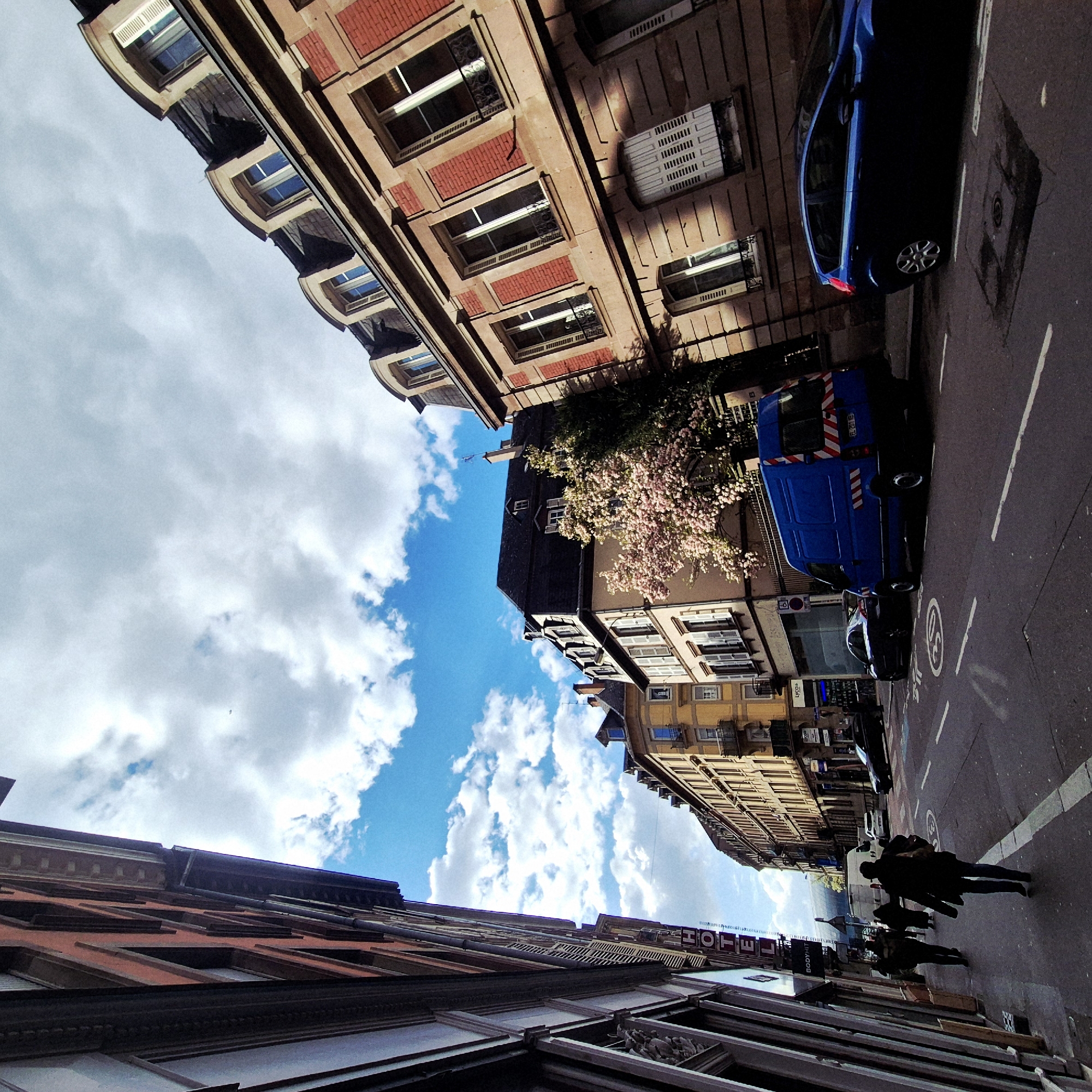 The image depicts a street view in Strasbourg, France, taken from a low angle looking up. Buildings line both sides of the street. The architecture features brickwork and windows with shutters. A blooming tree adds a touch of nature to the urban landscape. A blue van and a car are parked along the right side of the street. People are walking on the sidewalk on the right. A sign with the word "HOTEL" is visible. The sky is blue with white clouds.
