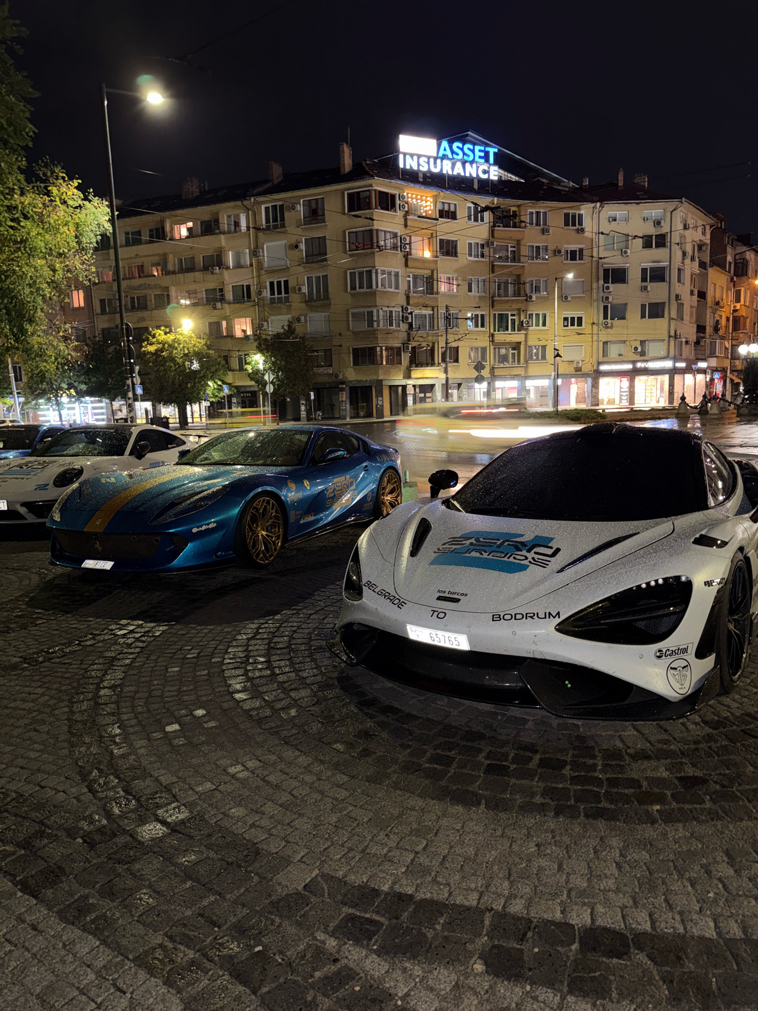 The image captures a night scene in Sofia, Bulgaria, with a row of luxury cars parked on a cobblestone street. In the foreground, a white McLaren sports car is prominently displayed, its hood wet from recent rain, adorned with decals that include "ZERO SURDS," "los turcos," "TO BODRUM," and a "Castrol" logo. To its left, a vibrant blue Ferrari is parked, also glistening from the rain, featuring gold rims and various sponsor decals. Further back, a white Porsche is visible, partially obscured.
The background features a multi-story apartment building with numerous lit windows, suggesting residential occupancy. The building's facade is typical of Soviet-era architecture. A prominent neon sign on the rooftop of the building reads "ASSET INSURANCE." Streetlights illuminate the scene, casting a warm glow on the wet pavement and the cars. The overall atmosphere is one of urban nightlife, highlighting the juxtaposition of high-end vehicles against the backdrop of a residential city environment. The wet cobblestone streets and the reflections of lights suggest that it has recently rained or is still drizzling. No people are visible in the frame.